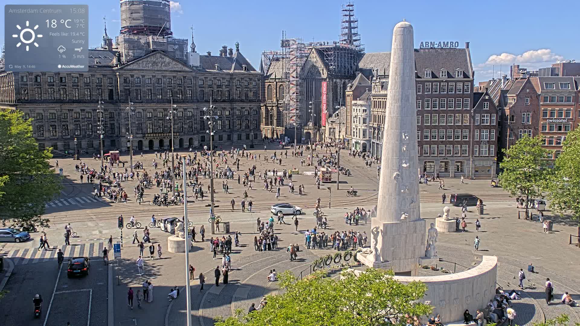 A sunny city square with a tall monument in the center, surrounded by buildings and numerous people, some sitting and others walking.