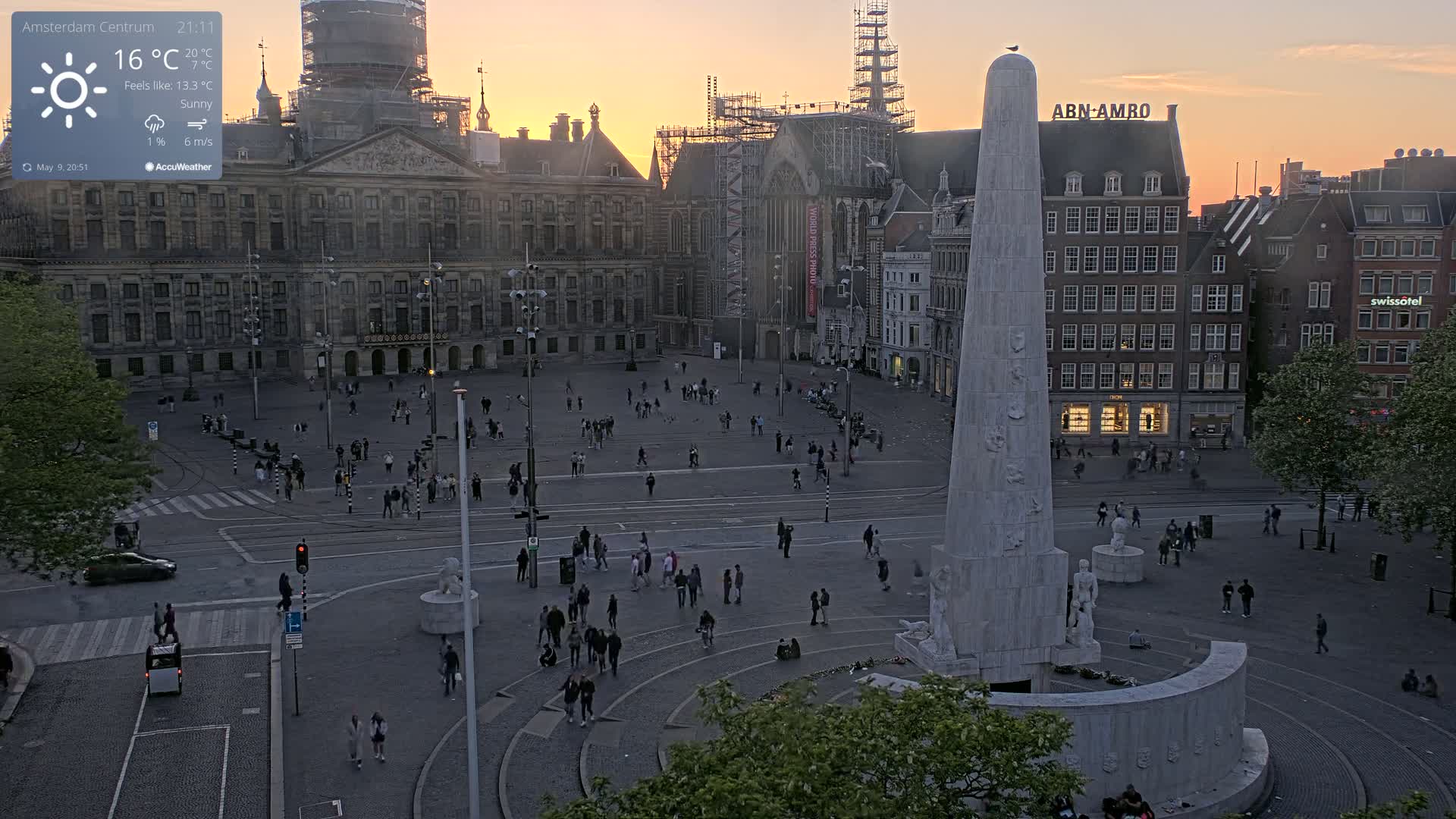 A large, mostly empty city square at sunset, with a tall monument in the center and several buildings surrounding it, under sunny skies.
