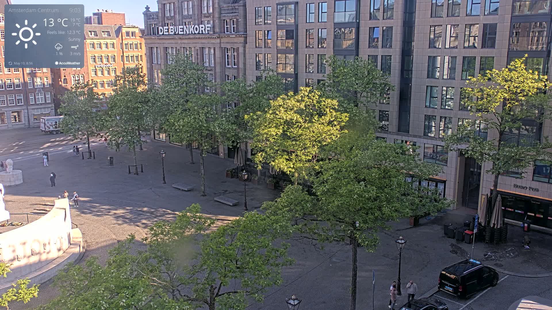 A sunny city square with several trees and a few pedestrians, under a partly cloudy sky.