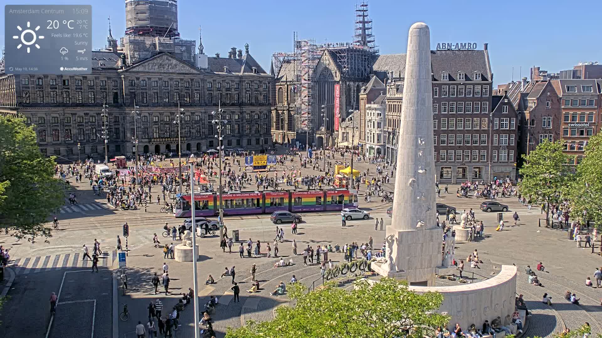 A sunny city square, filled with people, features a tall monument at its center and a rainbow-colored tram passing through.