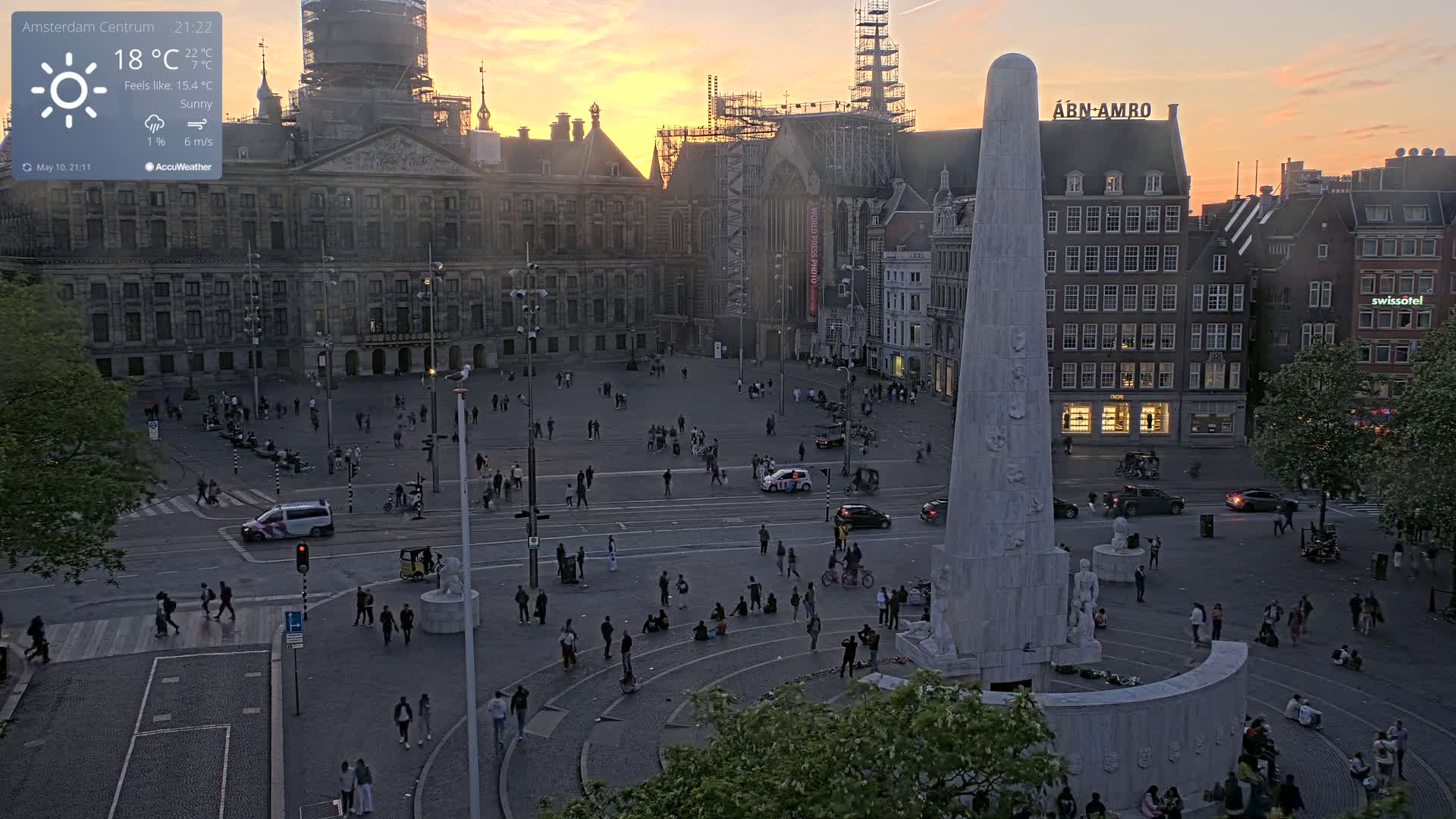 A large city square at sunset with many people scattered throughout, a tall monument in the center, and buildings surrounding the square.