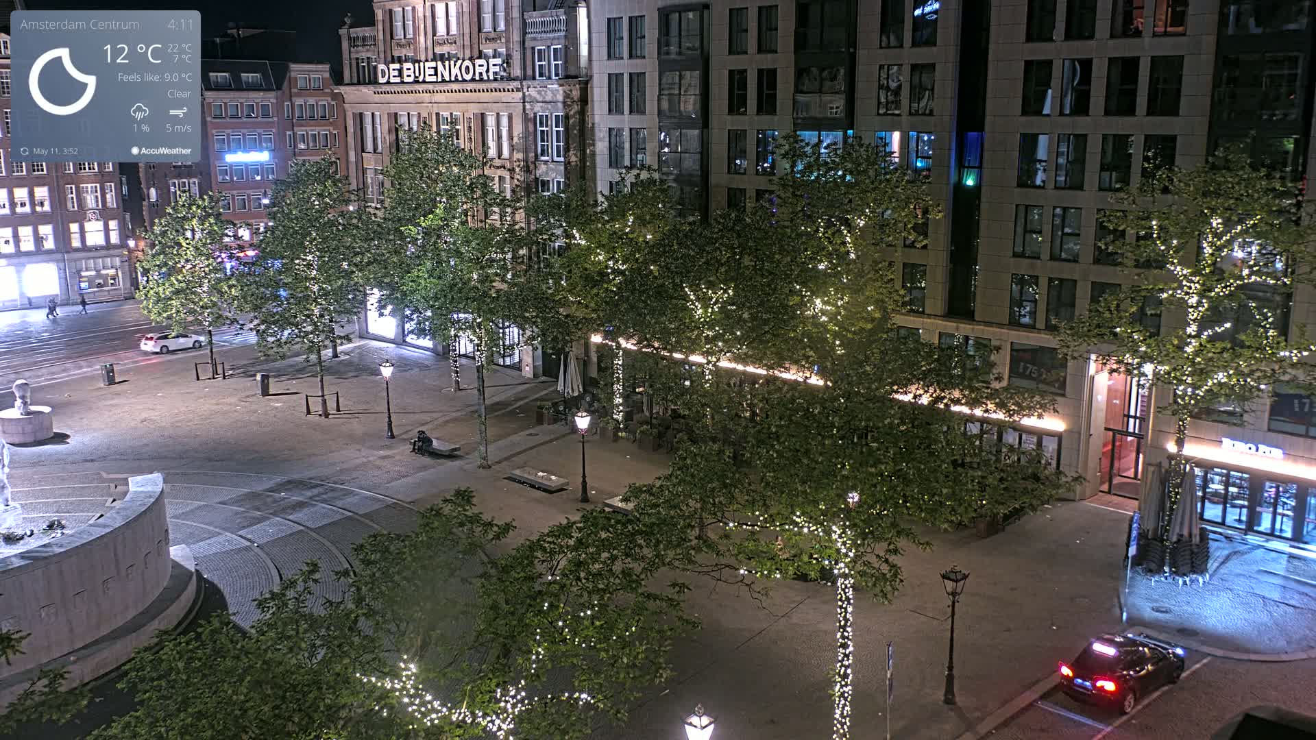 A nighttime view of a city square, illuminated by streetlights and strings of lights on trees, with a few pedestrians and vehicles present under clear skies.