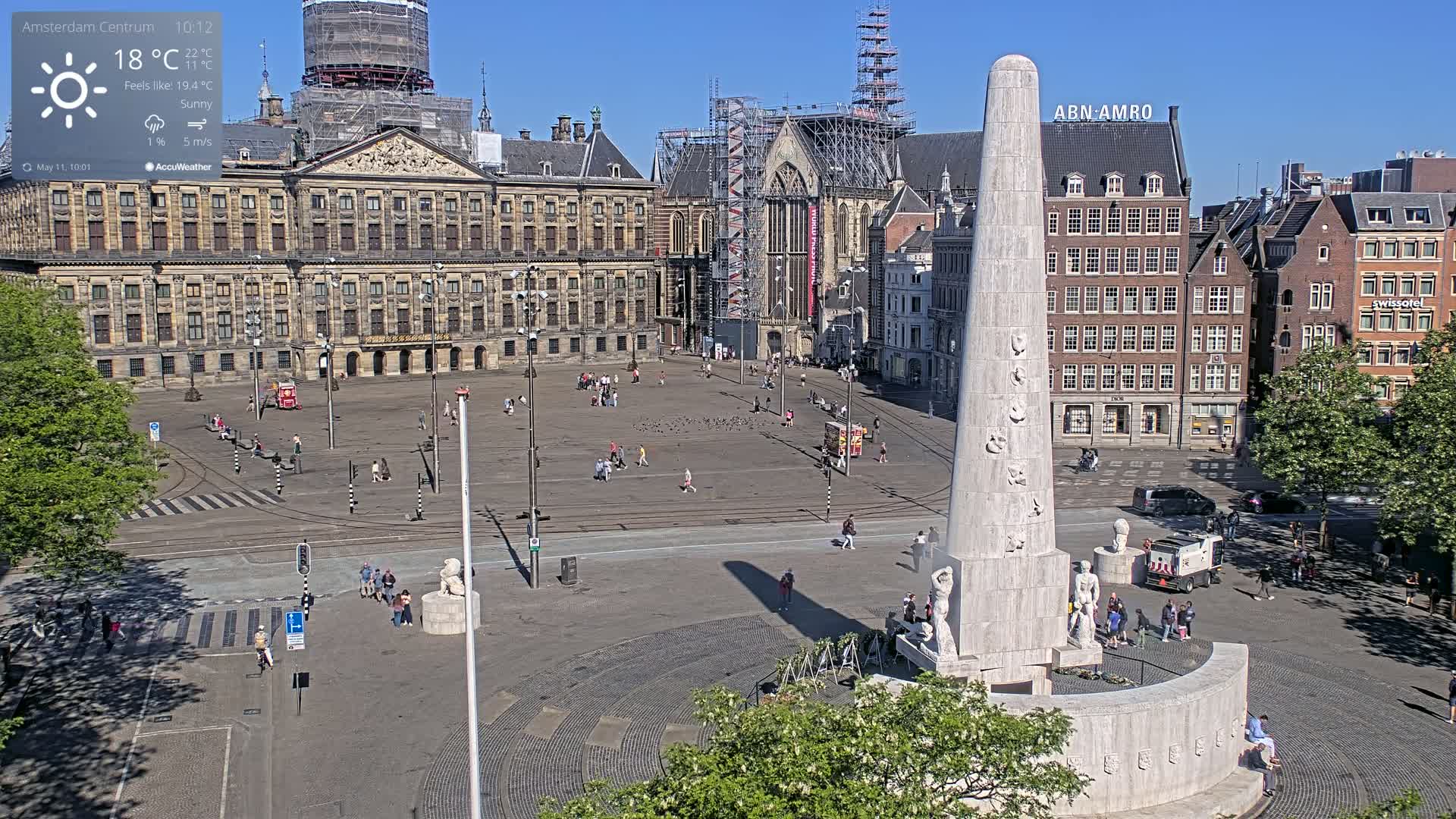 A sunny city square with a large monument at its center, surrounded by historical buildings and a few pedestrians.