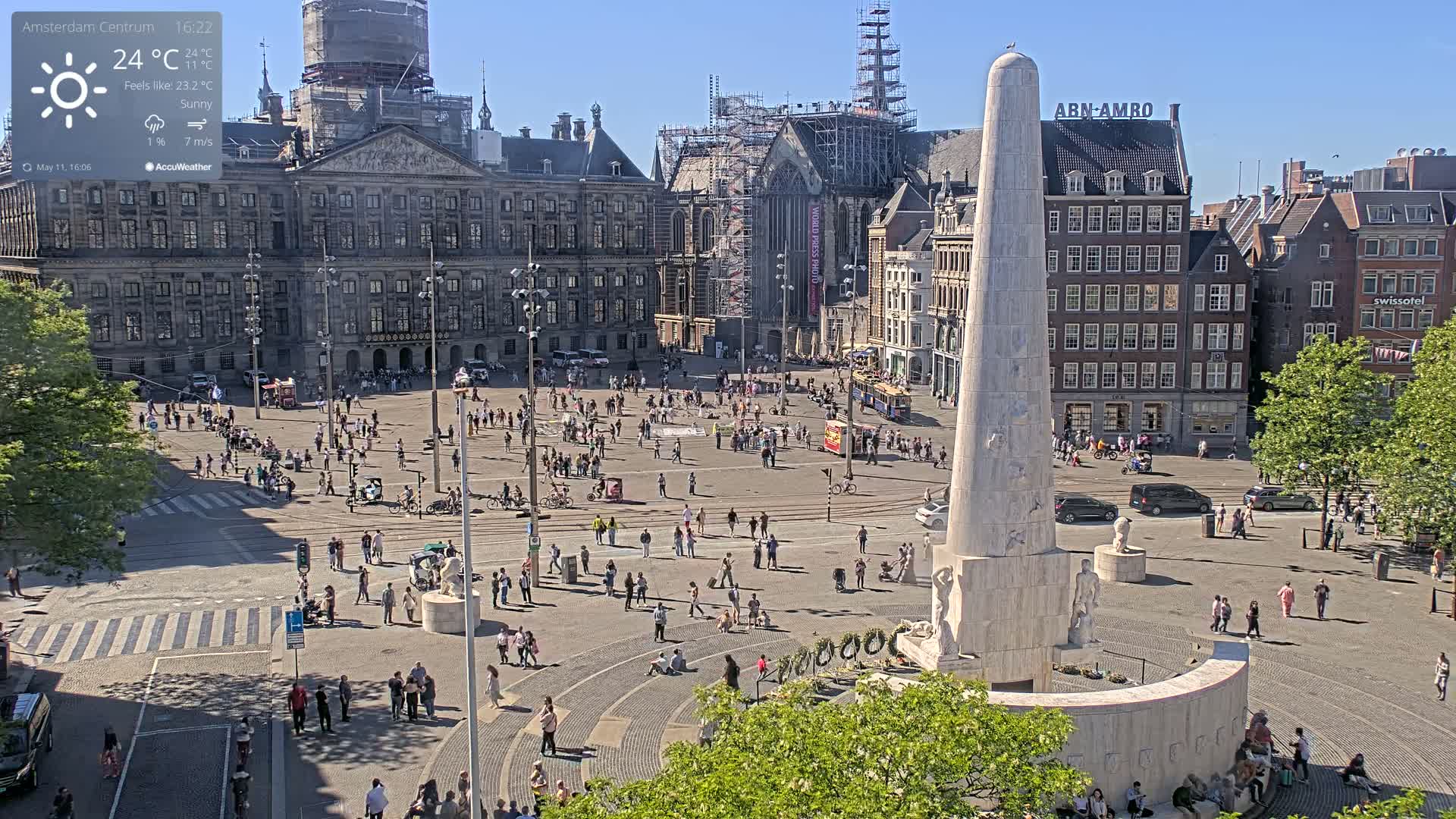 A sunny city square with a large monument, numerous people, and buildings surrounding it.
