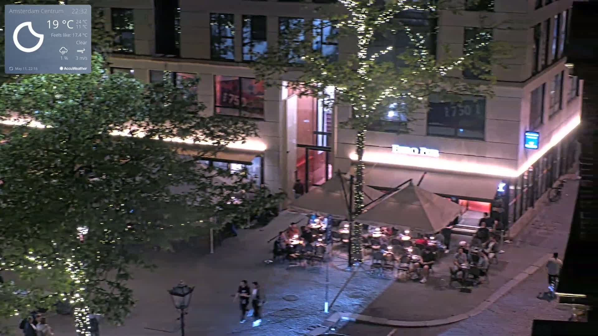 At night, under a clear sky with a gentle breeze, people are seated at outdoor tables in front of a building with lights illuminating the scene.