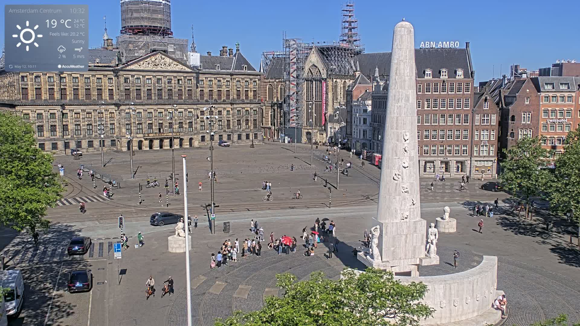 A sunny day in a large European city square shows a monument, a palace, surrounding buildings, and people walking around.