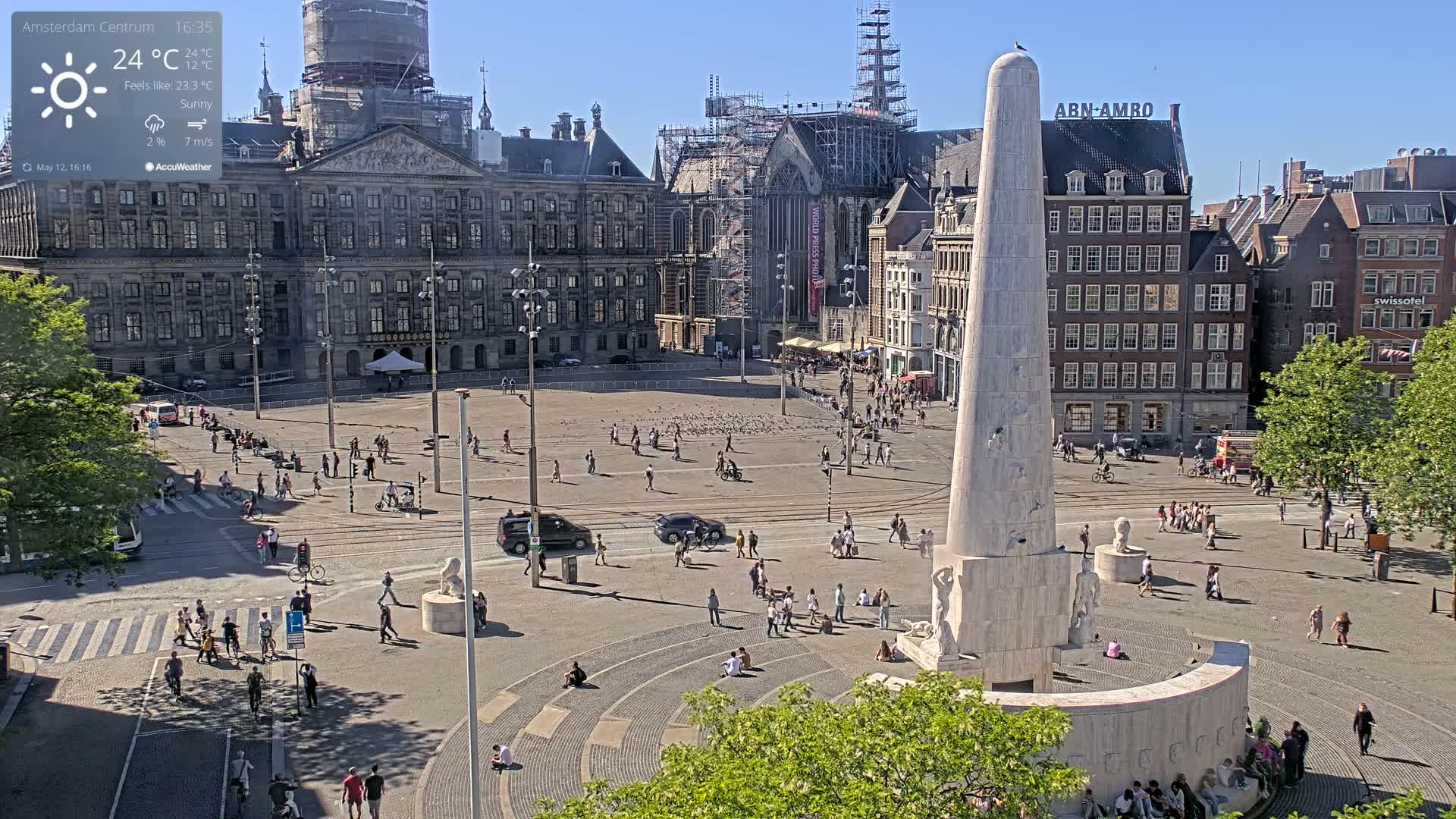 A sunny city square with a large monument at its center, surrounded by historical buildings, including one under construction, with numerous people walking and sitting.
