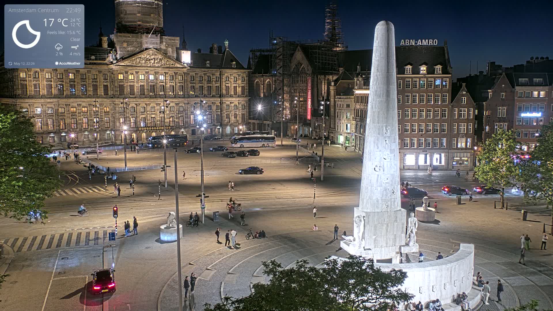 A nighttime, clear view of a city square with a tall monument at its center, surrounded by buildings and a few people and cars.