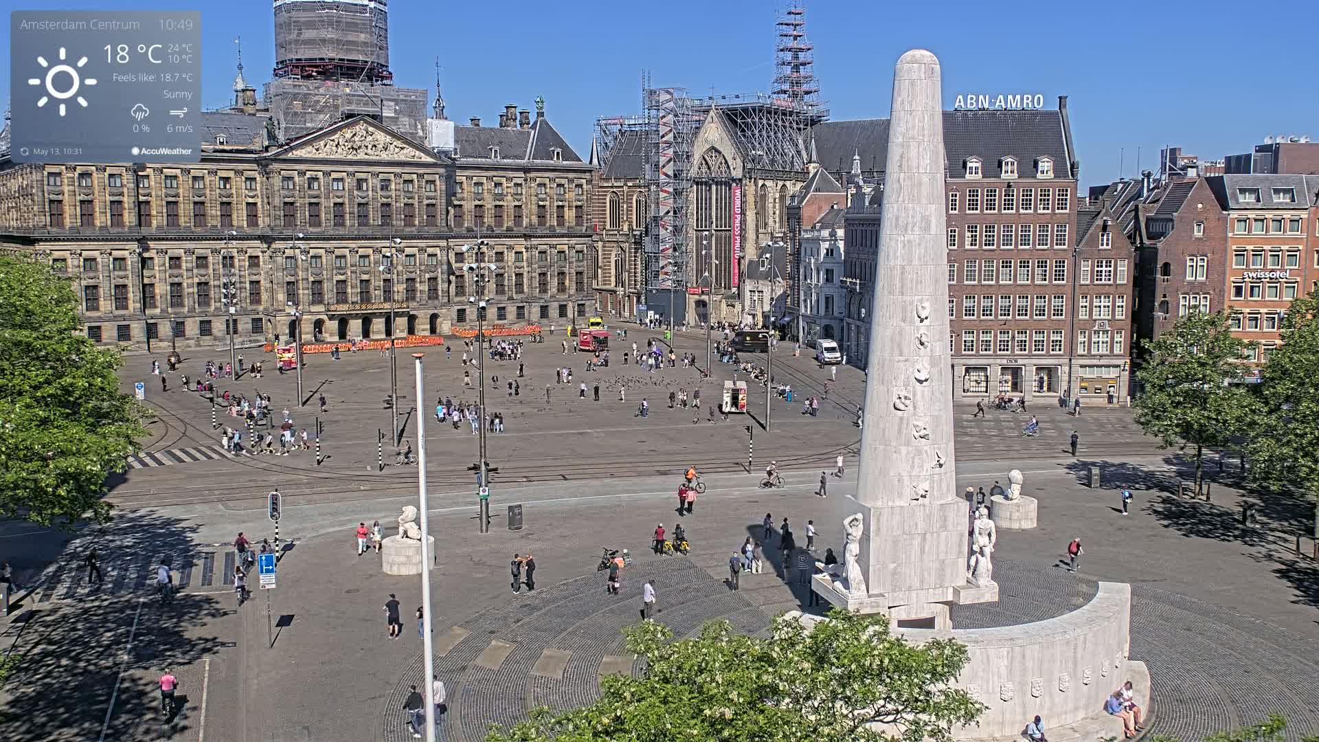 A sunny city square with a large monument at its center, surrounded by historical buildings and people walking and cycling.