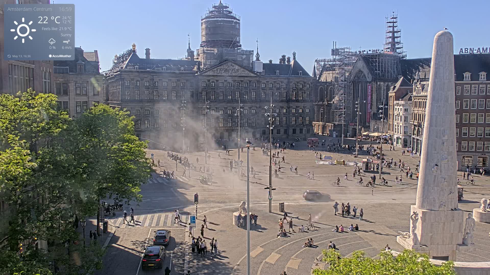 A sunny, hazy day in a large city square with a palace and monument, numerous people, and some construction scaffolding visible.
