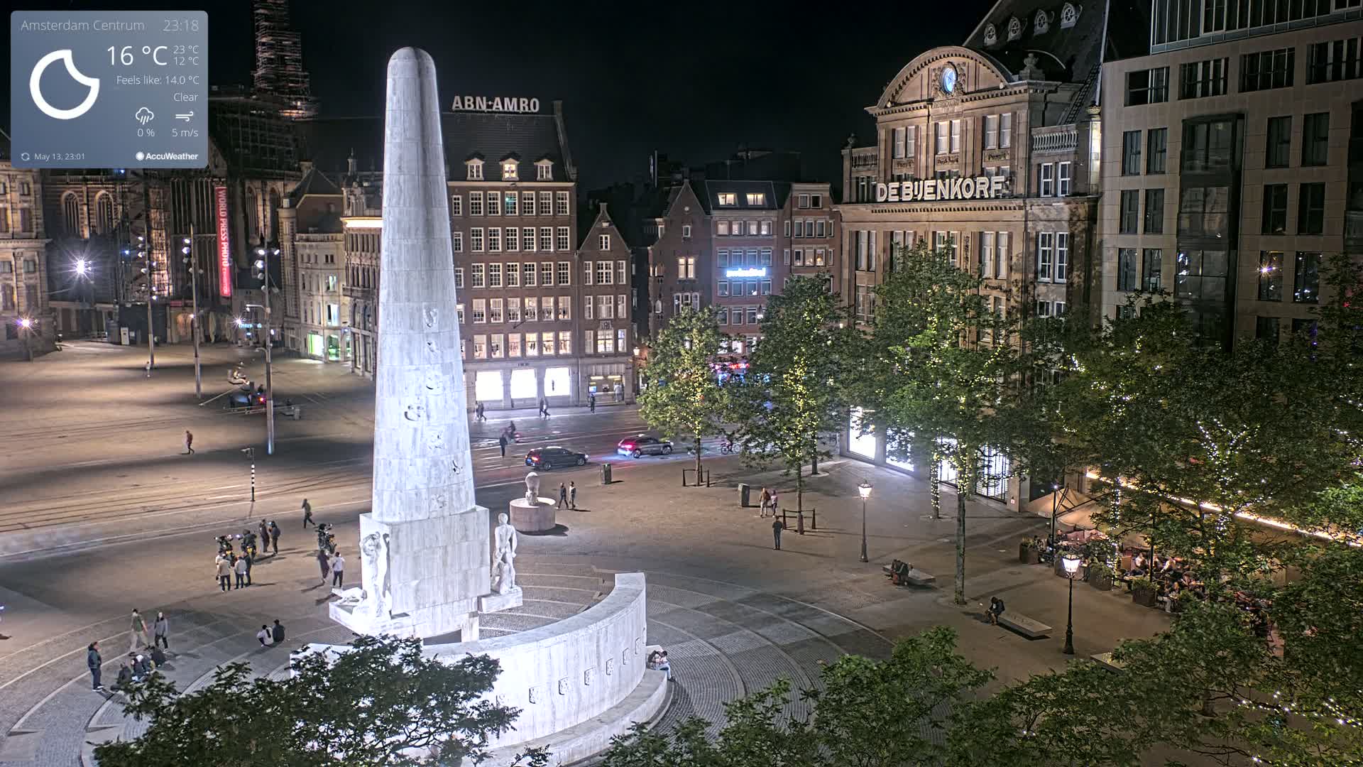 A nighttime, clear-weather view of a city square centered around a large monument, surrounded by buildings and trees with some string lights.