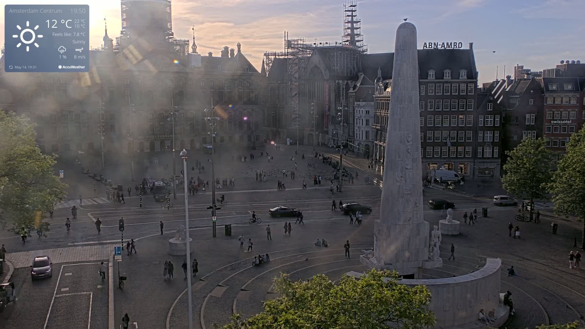 A hazy, sunny afternoon scene overlooking a city square with a tall monument at its center, surrounded by pedestrians and vehicles.