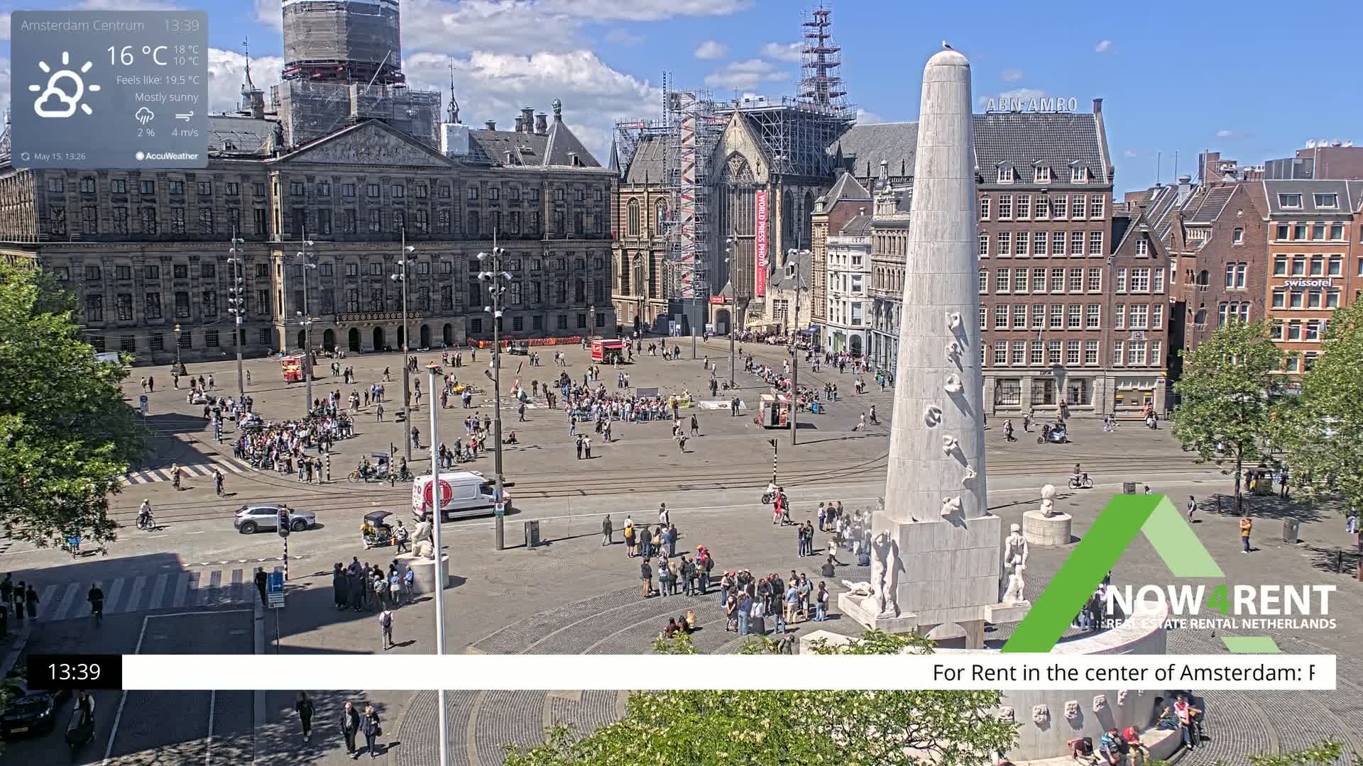 A mostly sunny city square with a large monument, numerous people, and surrounding buildings under a partly cloudy sky.