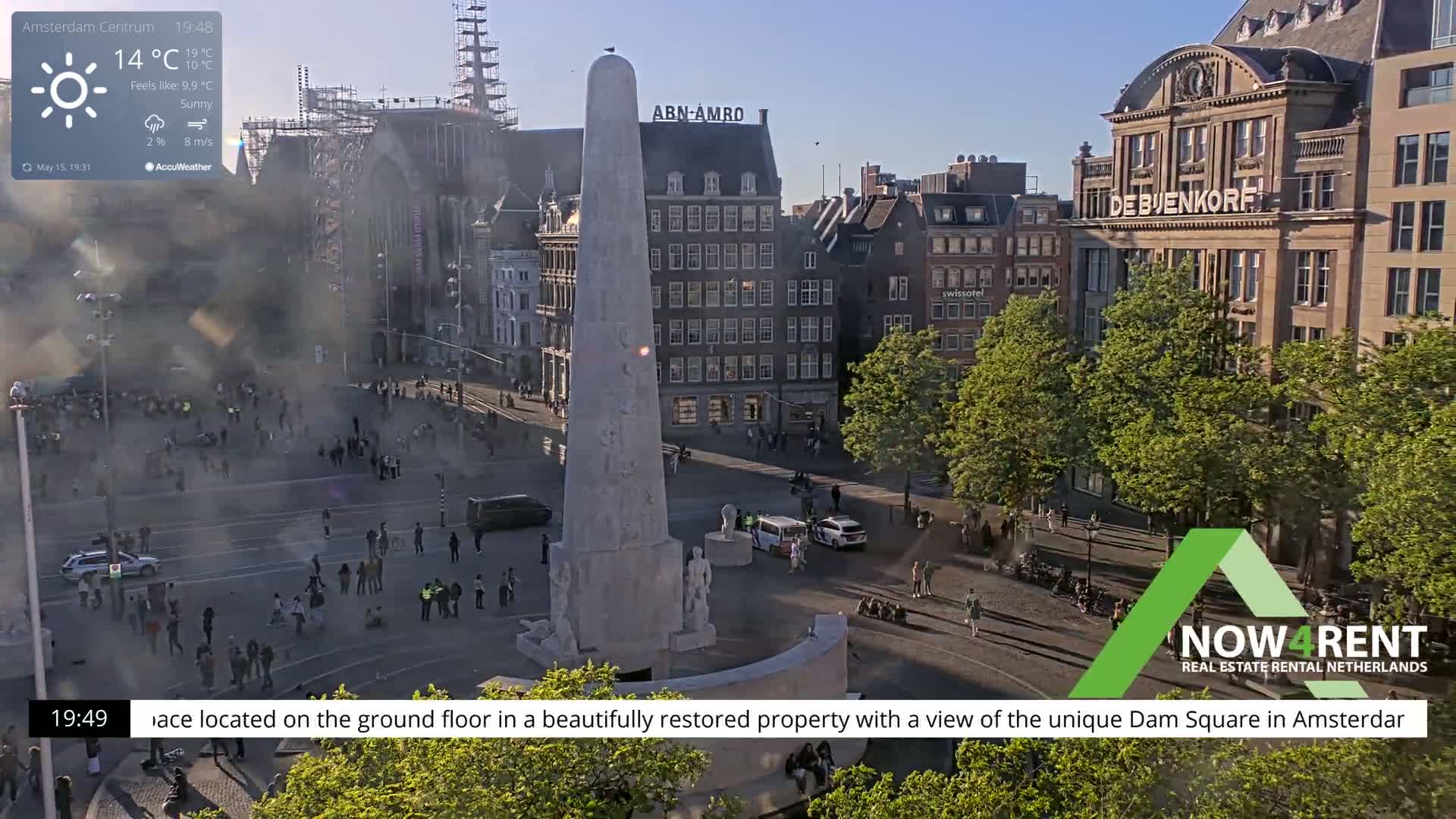 A sunny Dam Square in Amsterdam is viewed from above, showing a monument, surrounding buildings, and people walking around.