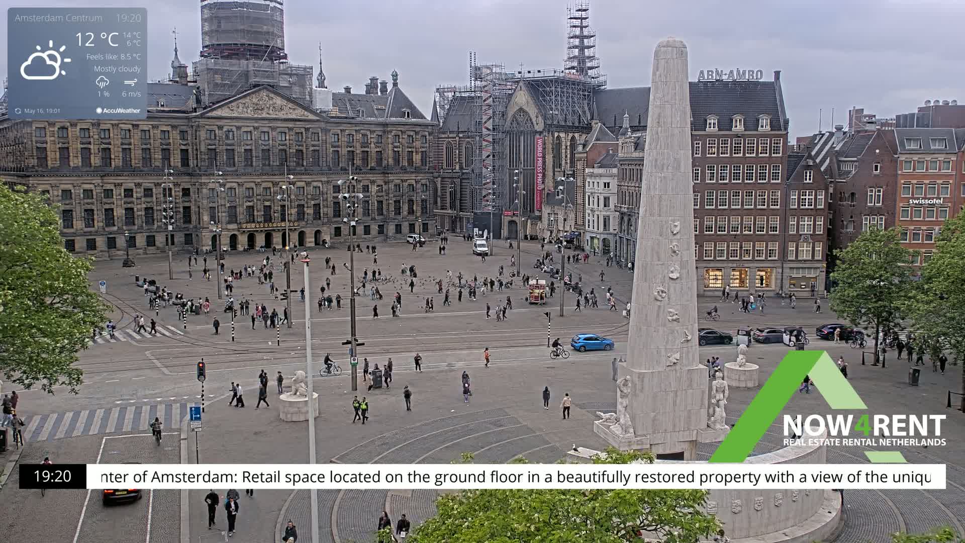 A mostly cloudy day in a large city square with a monument at its center, surrounded by historical buildings and pedestrians.