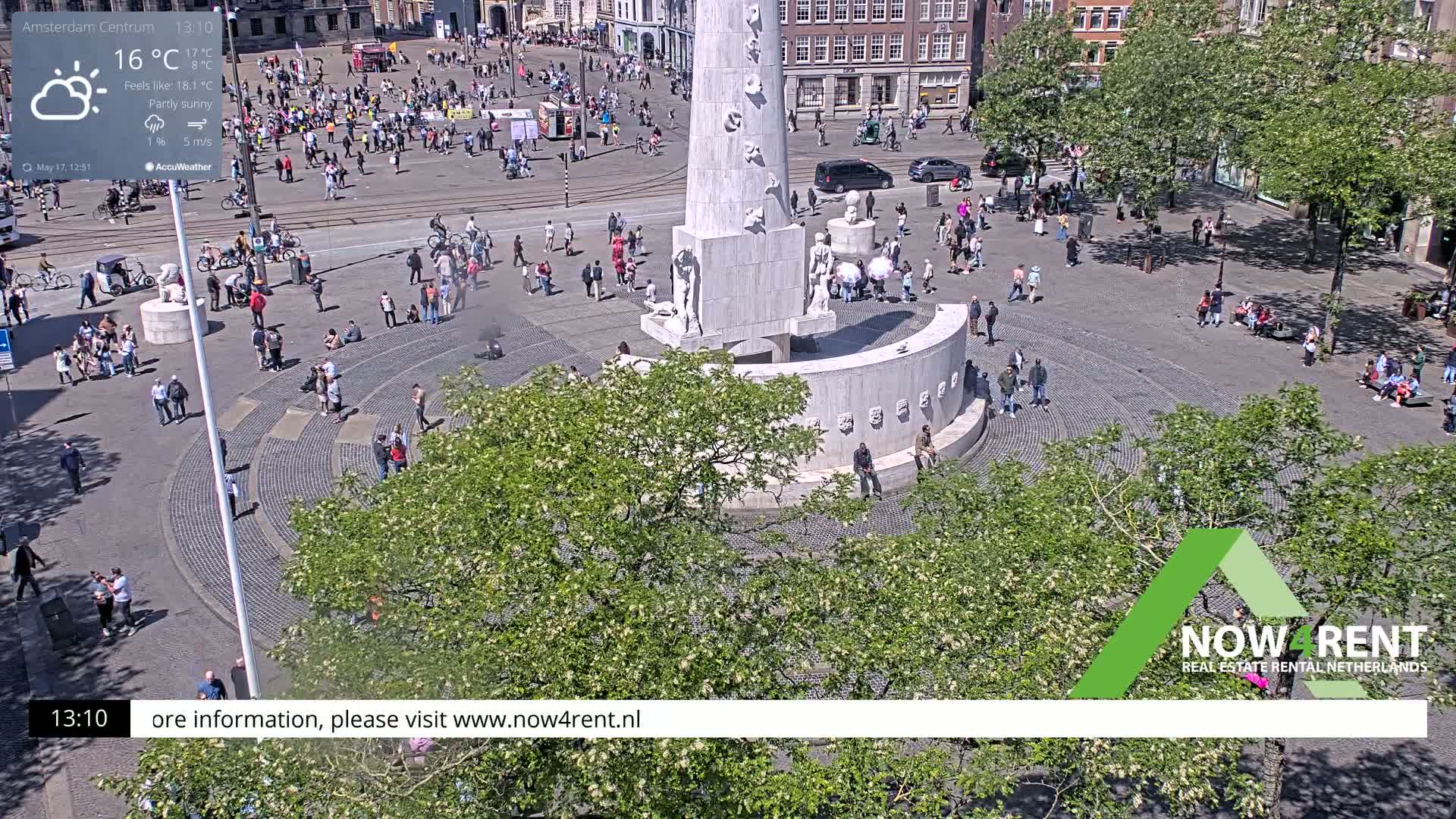 A partly sunny city square with a large monument in the center, surrounded by many people and trees.