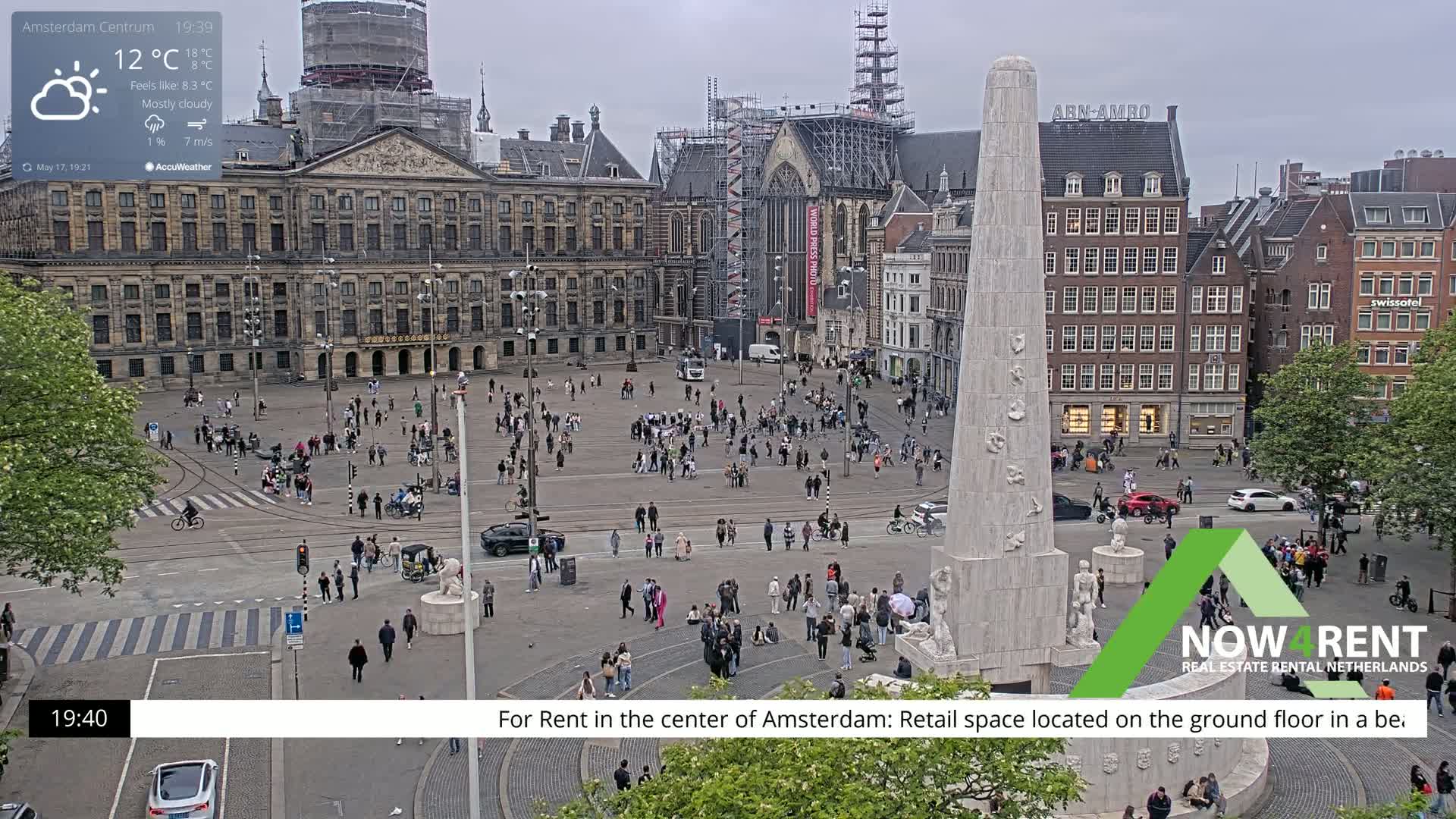 A mostly cloudy, cool day in a large European city square with many people, buildings, and a tall monument at its center.