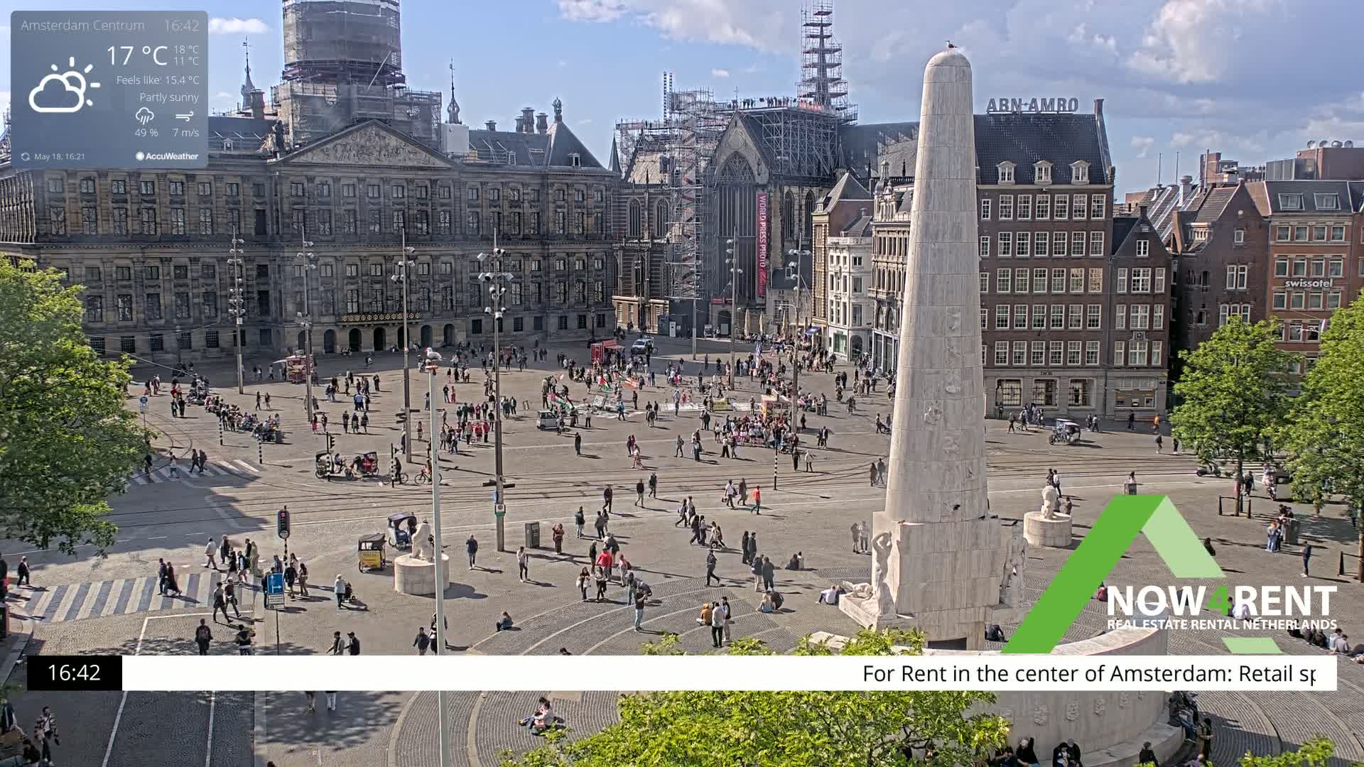 A partly sunny city square with many people walking around, a large monument in the center, and several buildings surrounding it.