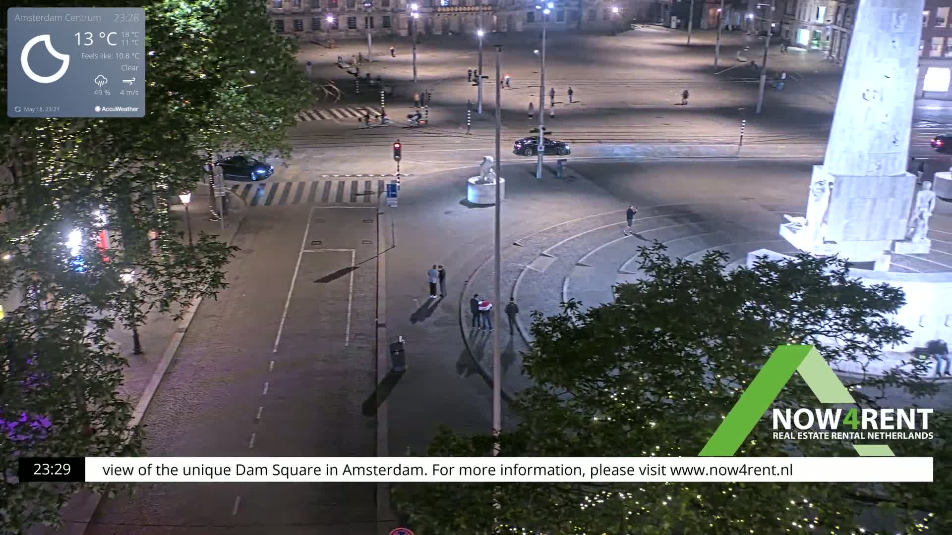 A nighttime, clear-weather view of a large, mostly empty city square with a monument and several people sparsely scattered throughout.
