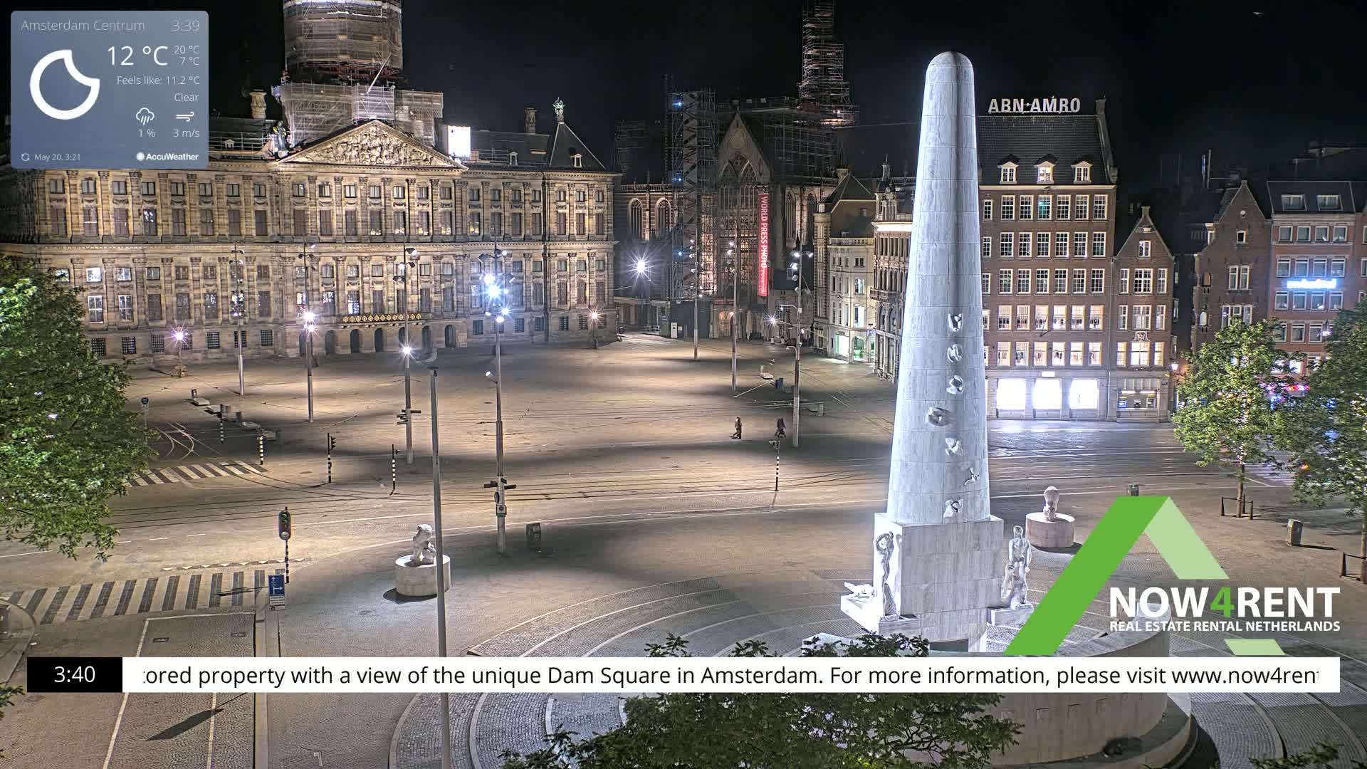 A nighttime, clear view of Dam Square in Amsterdam, featuring a tall monument in the foreground and various buildings illuminated around the square.