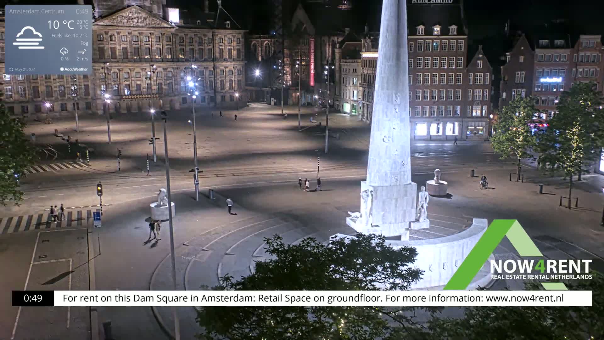 A nighttime, foggy view of a city square with a tall monument in the center, surrounded by buildings and a few pedestrians.