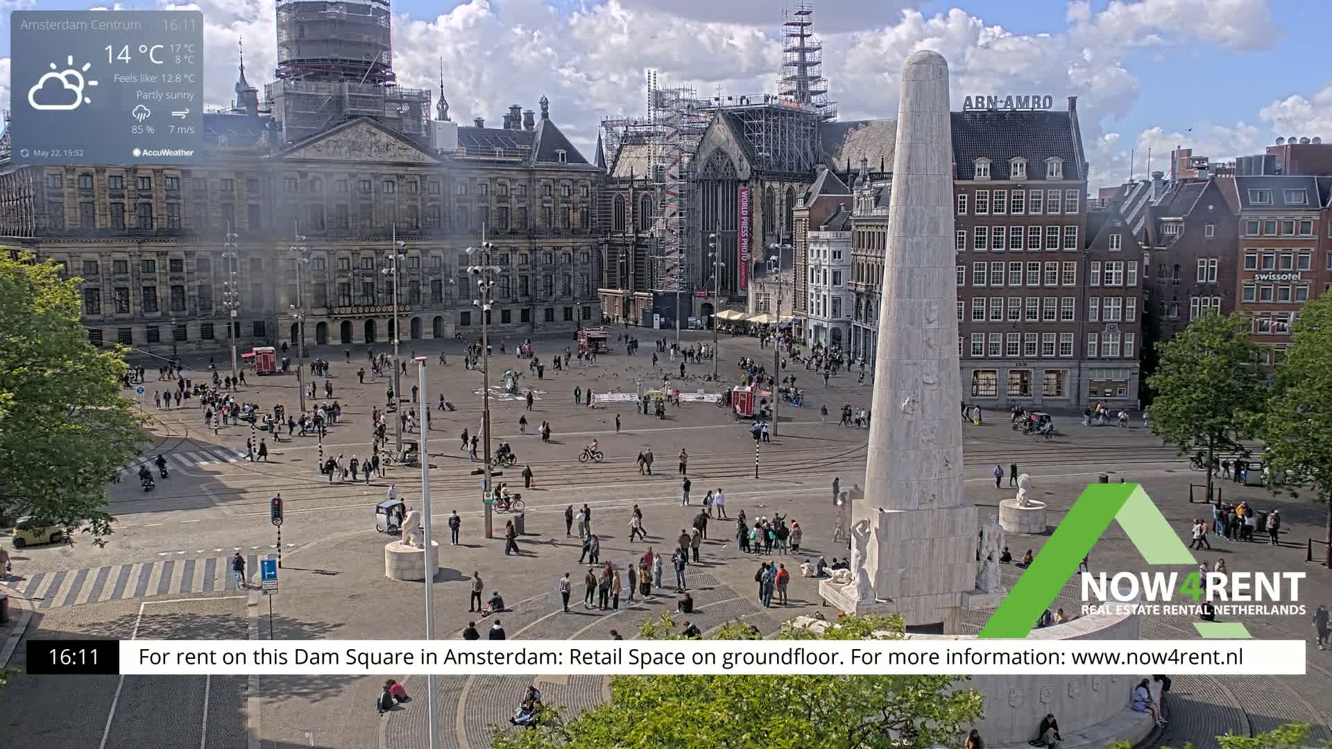 A partly sunny Dam Square in Amsterdam, with a monument in the foreground and many people walking around.