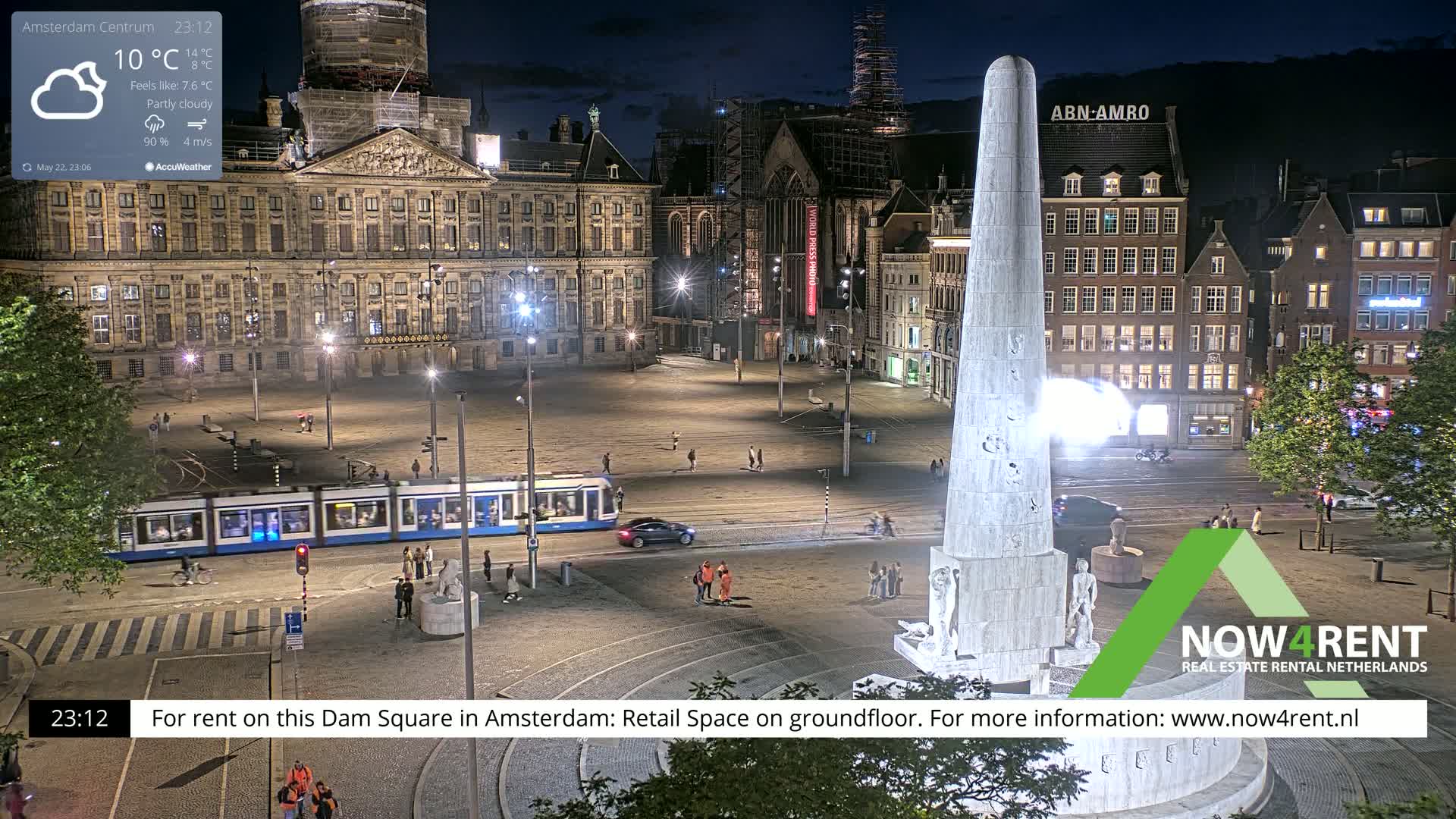 A nighttime view of a city square with a monument in the center, a tram, and several buildings, under partly cloudy skies.