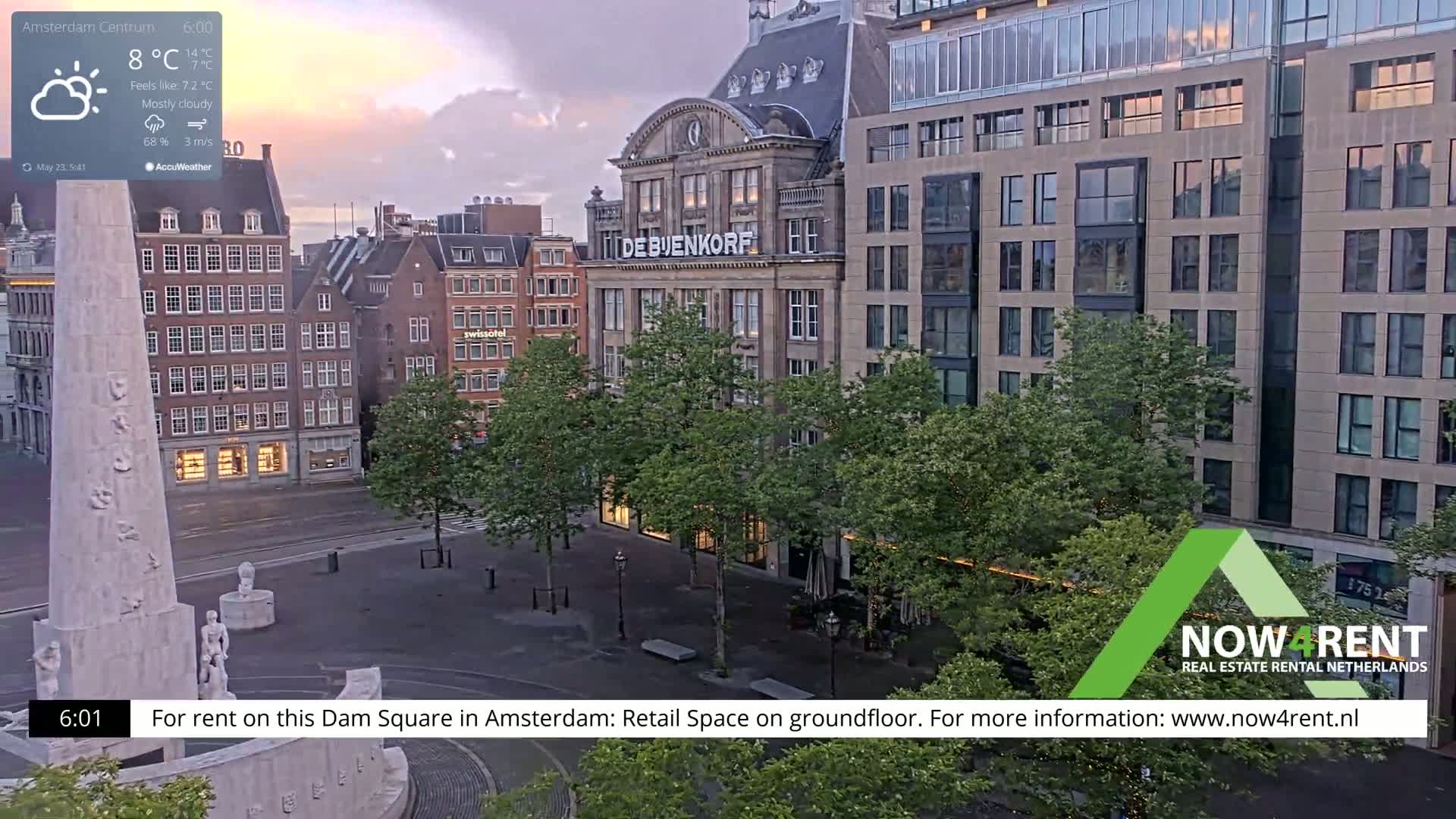 A mostly cloudy sunrise illuminates a city square with a monument, trees, and several buildings.