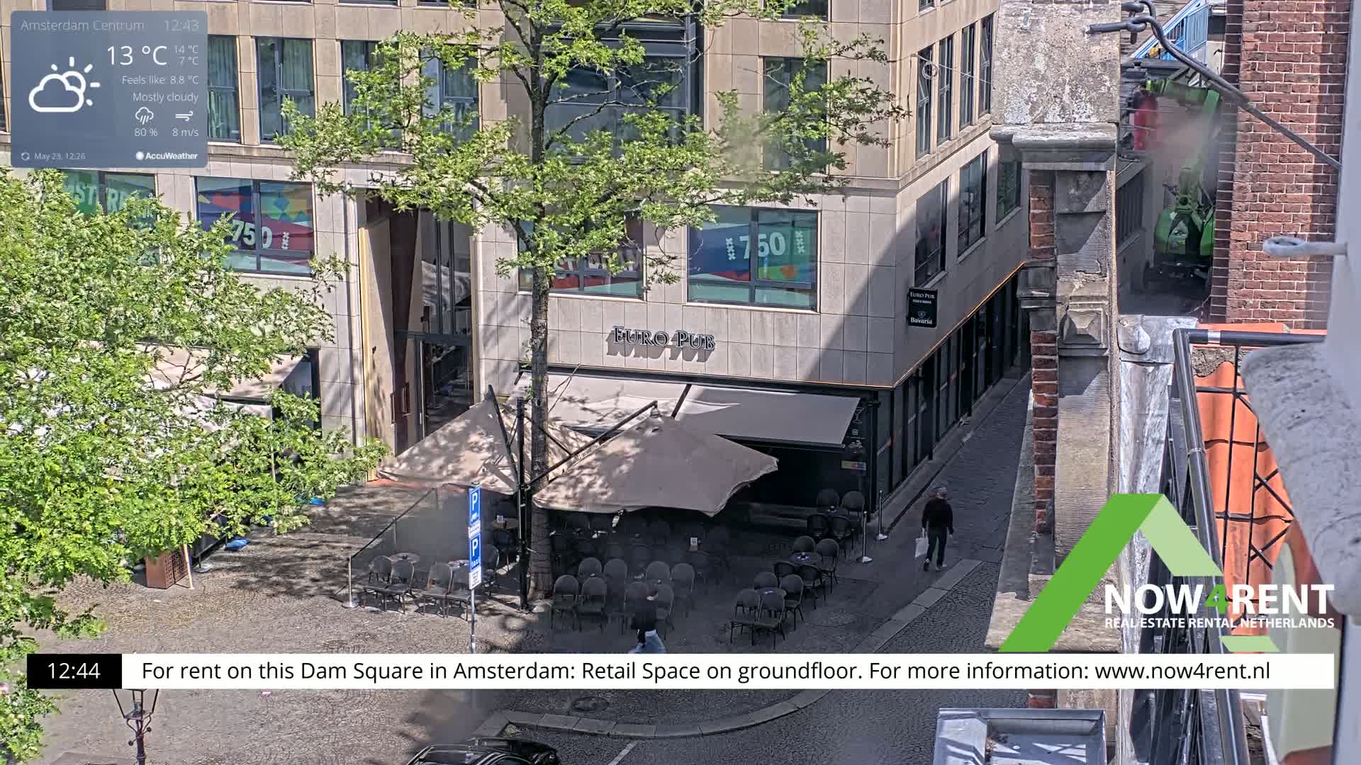 A mostly cloudy, sunny day shows an outdoor cafe with a few patrons seated at tables under umbrellas, situated beside a building with large windows.