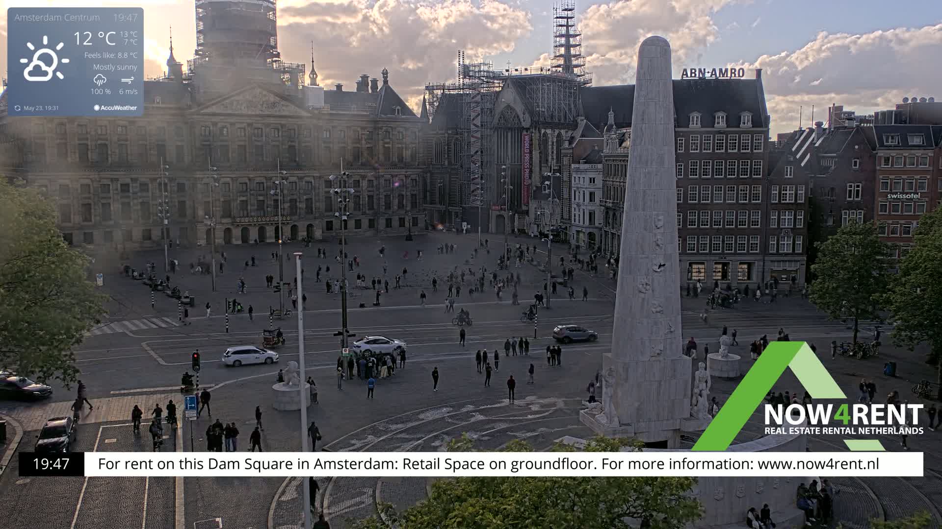 A mostly sunny city square with a large monument and surrounding buildings, under a partly cloudy sky.