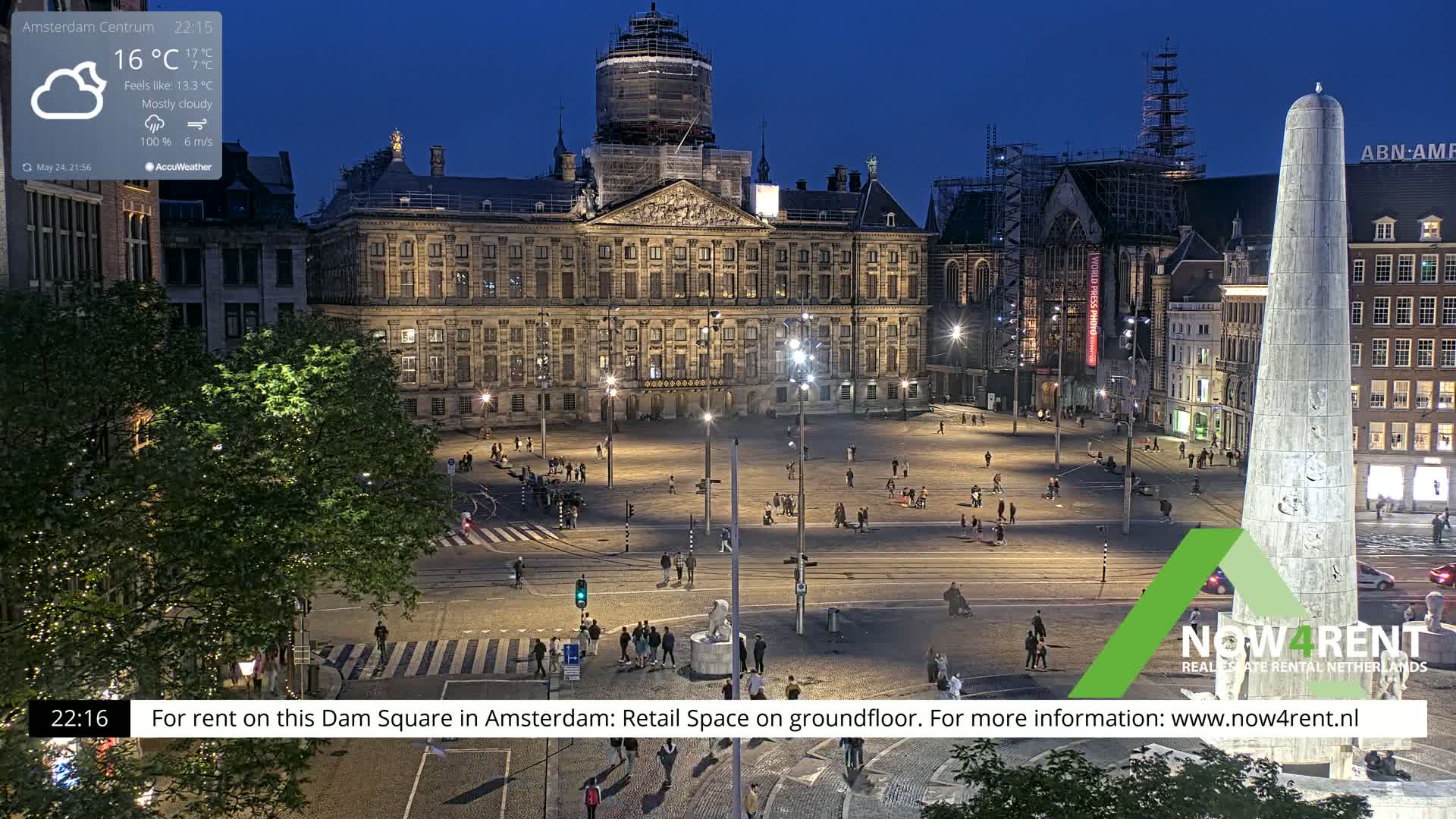 A mostly cloudy night scene shows a large square in a city with a palace, a monument, and several buildings illuminated, with a few people scattered throughout.