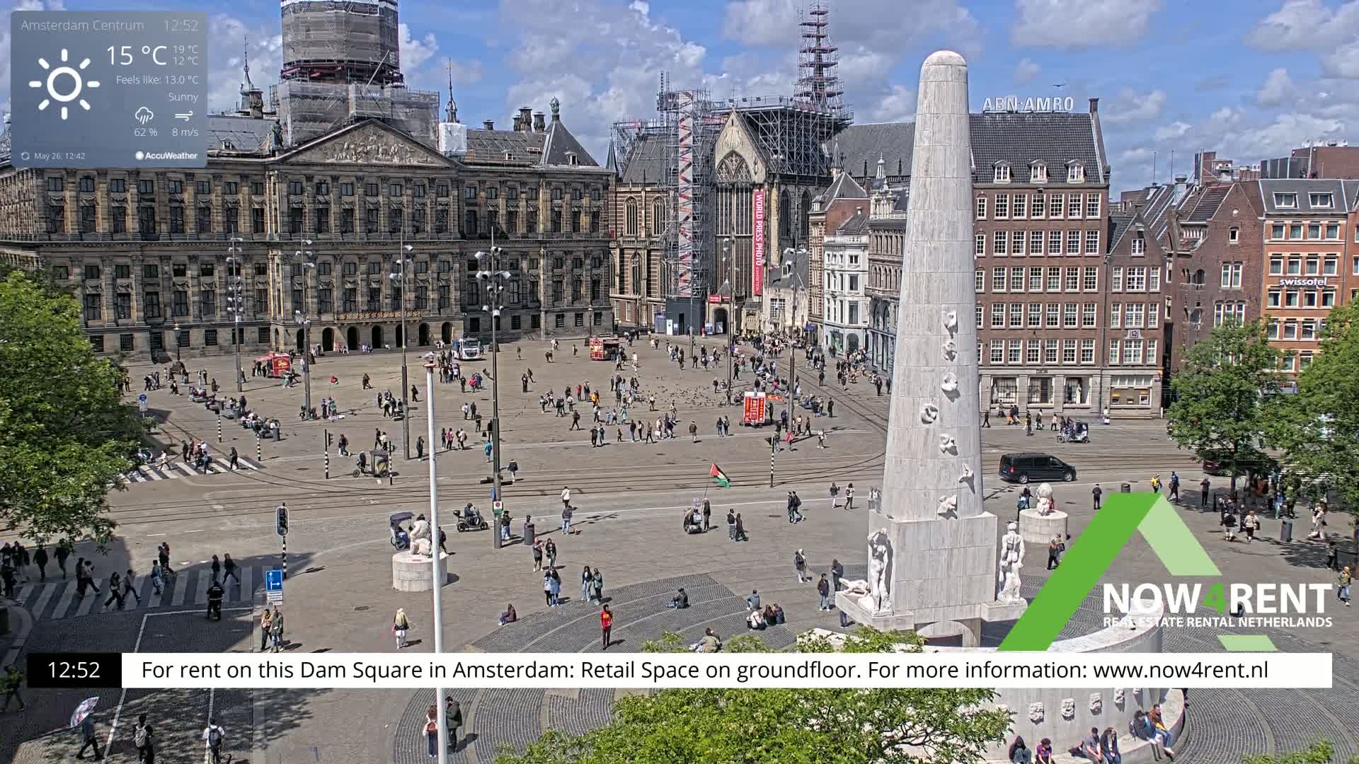 A sunny Dam Square in Amsterdam, with a large monument at the center, is populated by numerous people and features surrounding buildings, one of which is under construction.