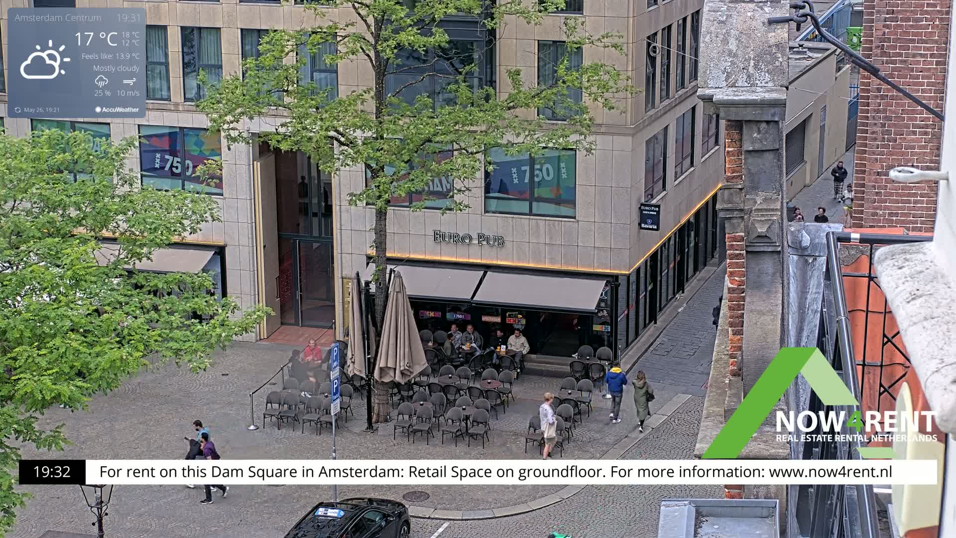 A mostly cloudy, cool evening scene shows an outdoor café with patrons seated at tables under an umbrella, situated on a cobblestone square in front of a building.