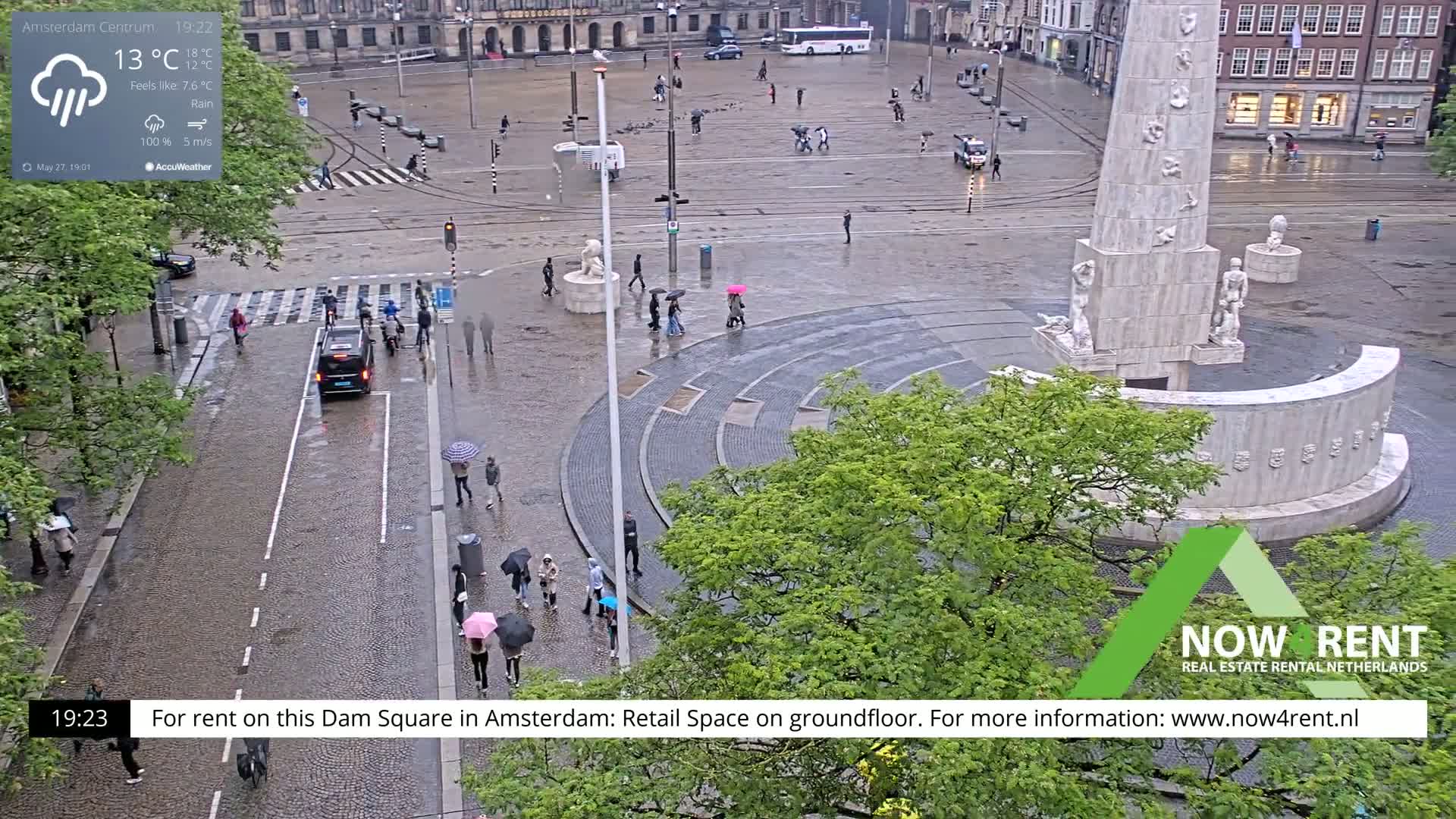 A rain-soaked Dam Square in Amsterdam shows pedestrians with umbrellas walking past a monument and across a plaza.