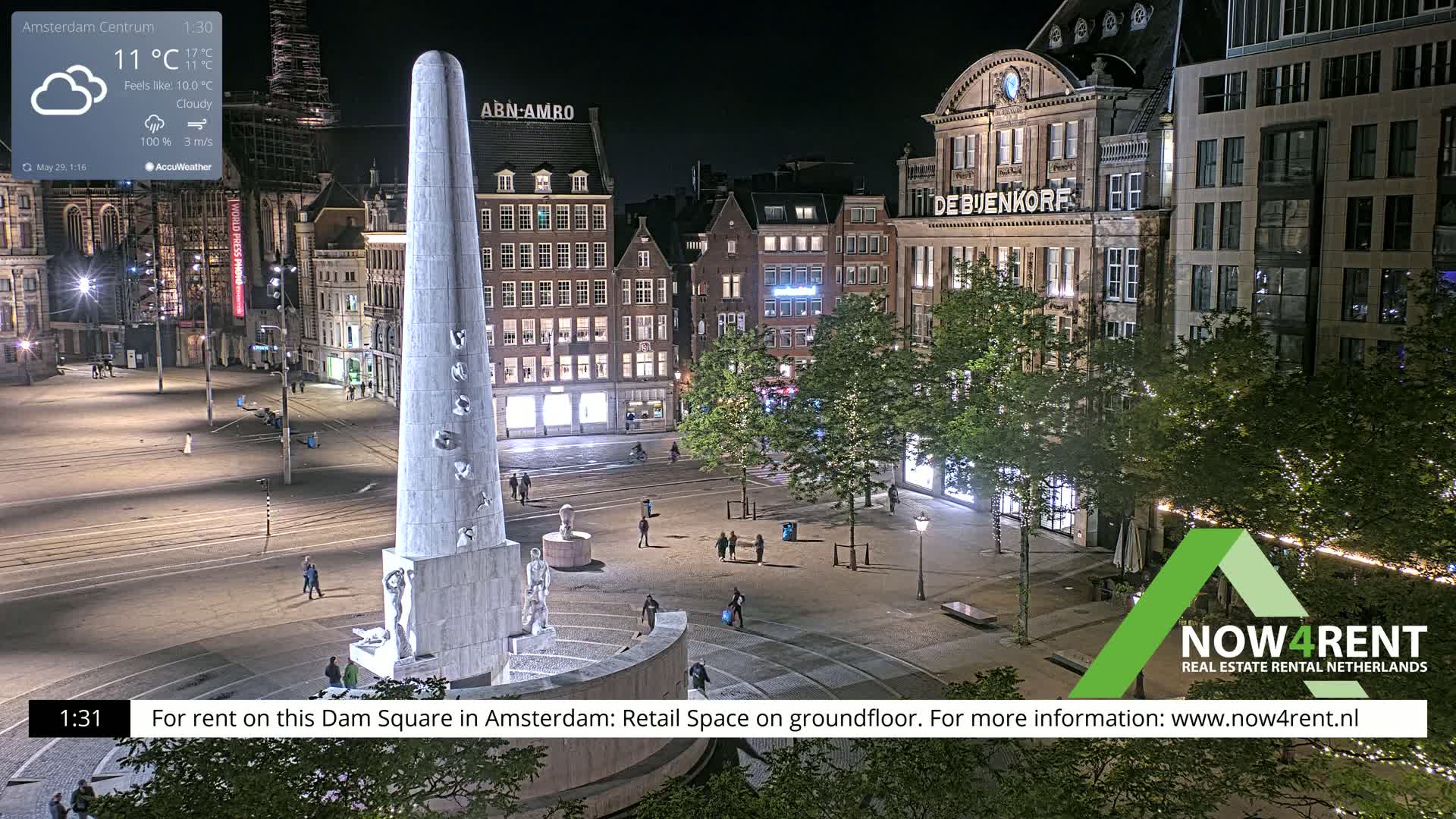 A nighttime view of Dam Square in Amsterdam, showing a monument, buildings, and people, under cloudy skies.