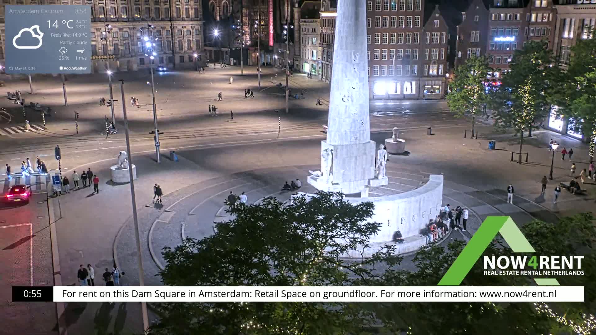 A nighttime, partly cloudy view of a large city square with a tall monument at its center, surrounded by people and buildings.