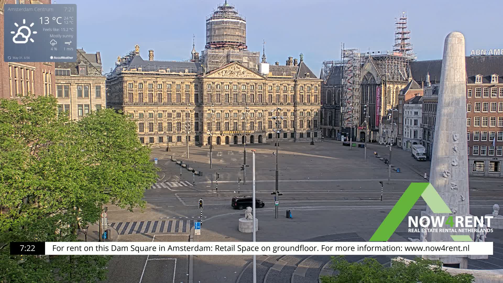 A mostly sunny, city square in Amsterdam, featuring a large palace under construction and a tall monument, is mostly empty with one car visible.
