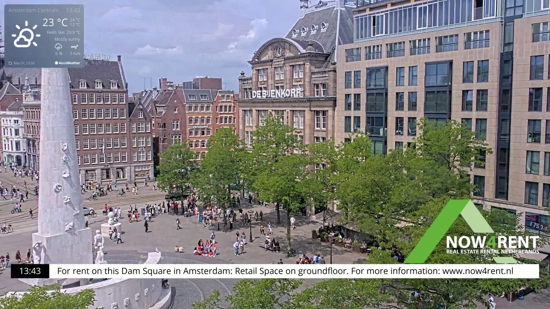 A mostly sunny view of Dam Square in Amsterdam, showing a monument,  buildings, and people walking around the square.