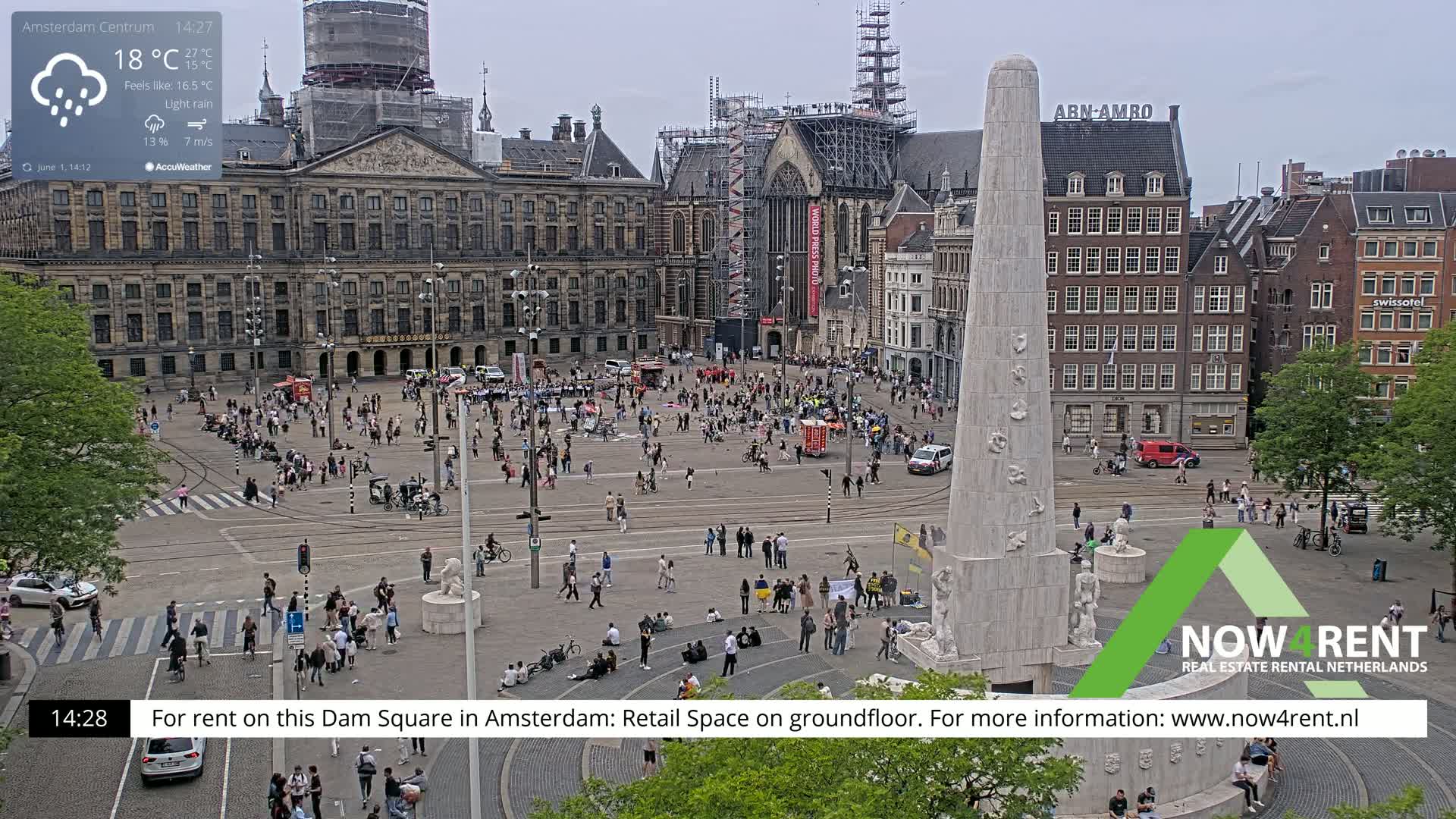 A bustling city square with a large monument, numerous pedestrians, and buildings under a lightly rainy sky.