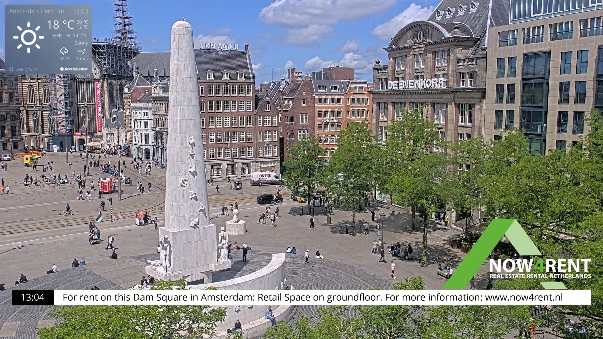 A sunny day in a city square, showing a monument surrounded by people and buildings, with green trees lining the edges.