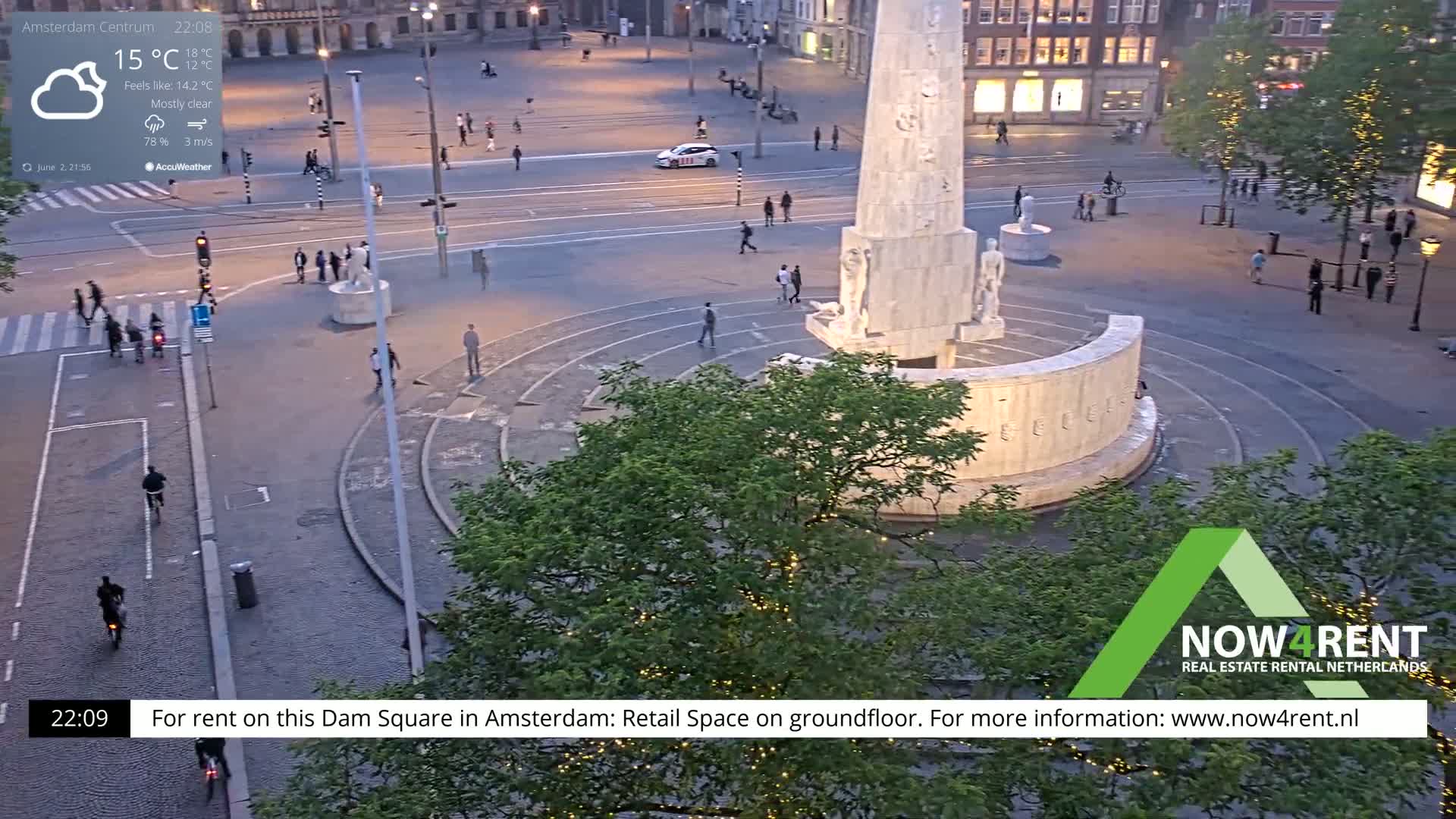 A mostly clear, cool evening shows a sparsely populated Dam Square in Amsterdam, centered on a large monument surrounded by trees and streetlights.