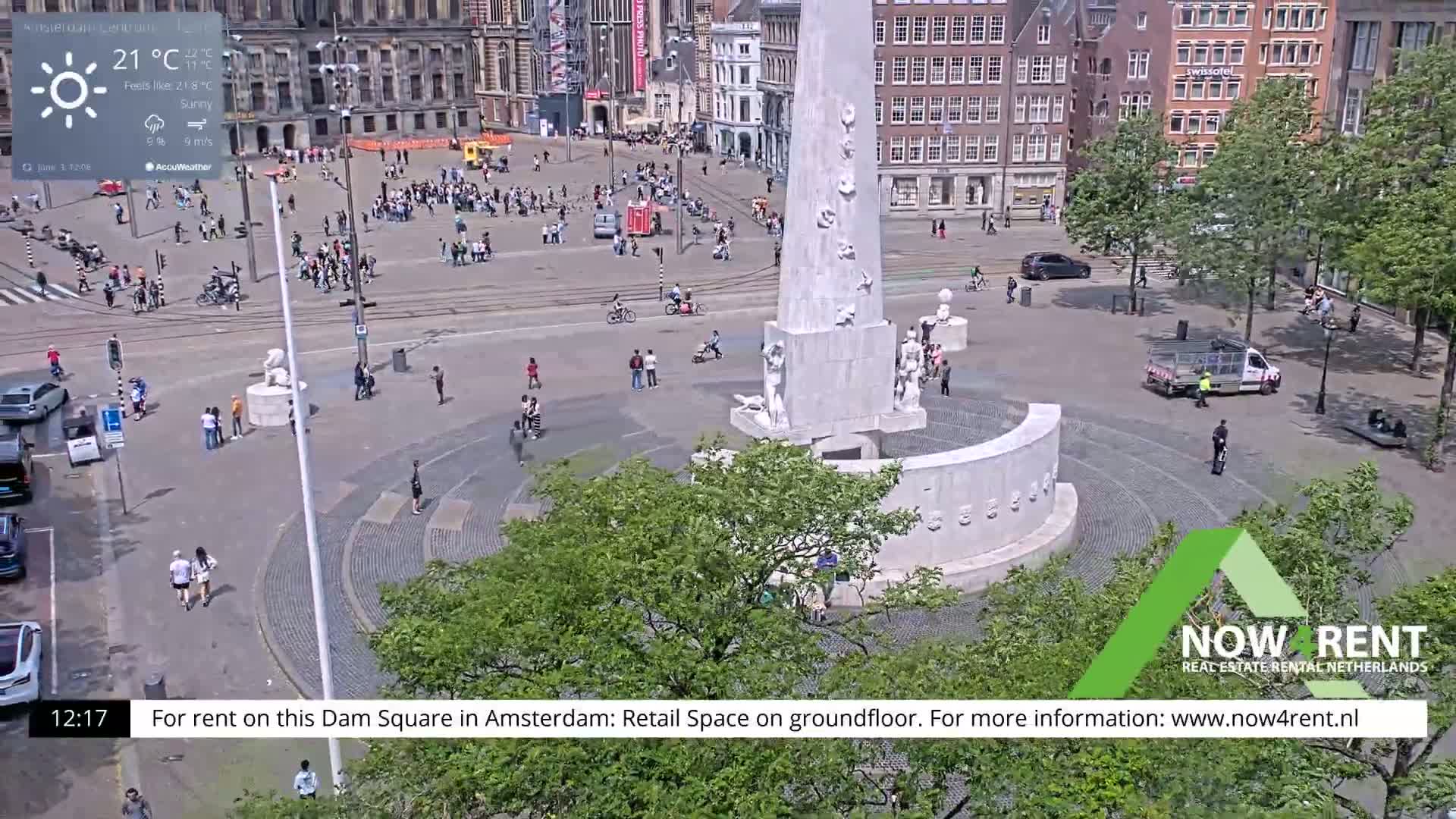 A sunny day in a city square with a large monument in the center, surrounded by people and buildings.