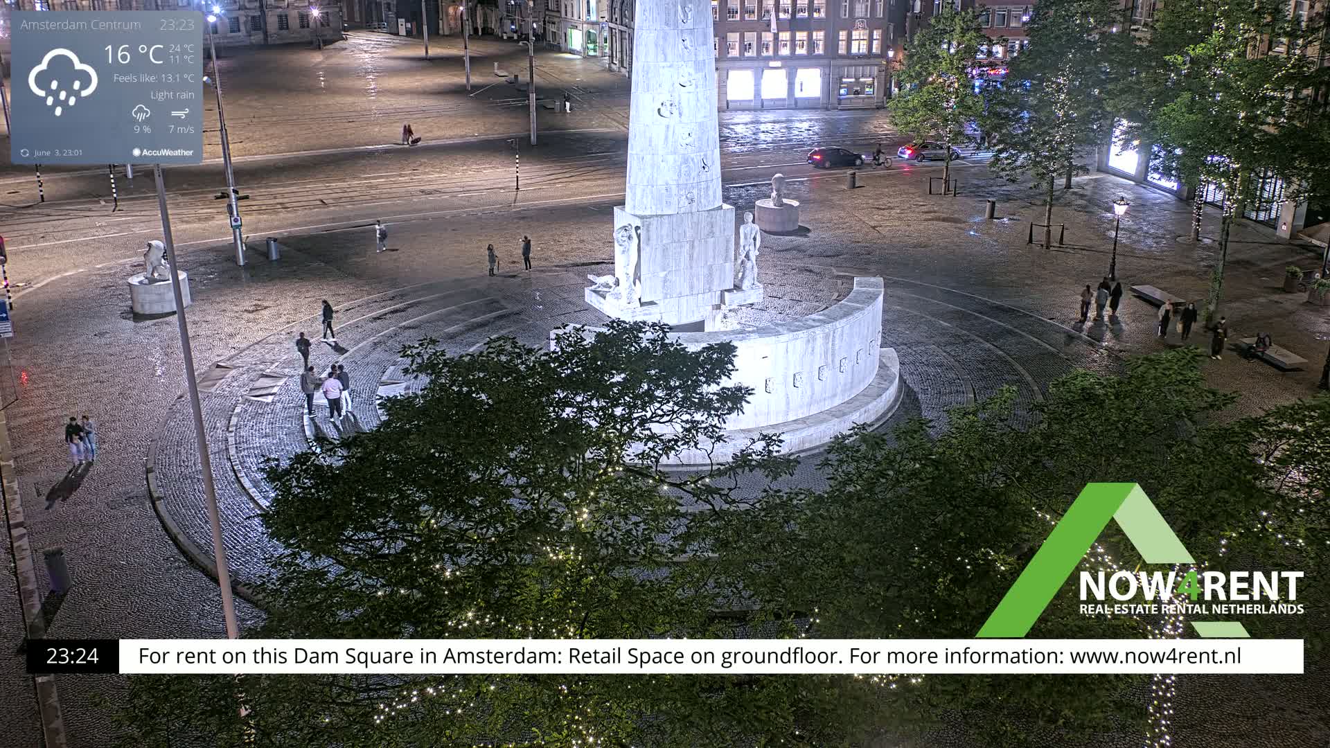 A nighttime, lightly raining view of a large, circular plaza centered around a tall stone monument, with several people walking around.