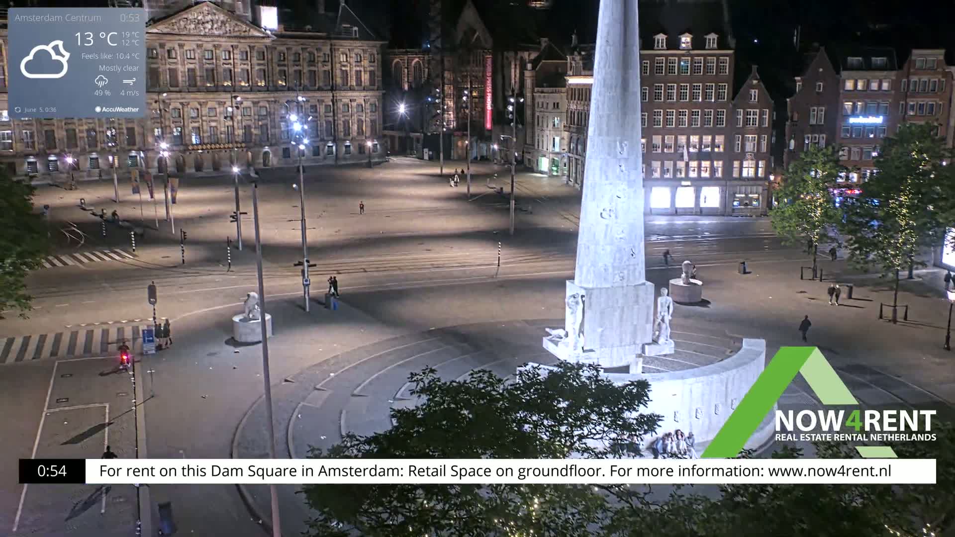 A mostly clear night scene shows a sparsely populated Dam Square in Amsterdam, centered on a large monument, with surrounding buildings and streets illuminated.