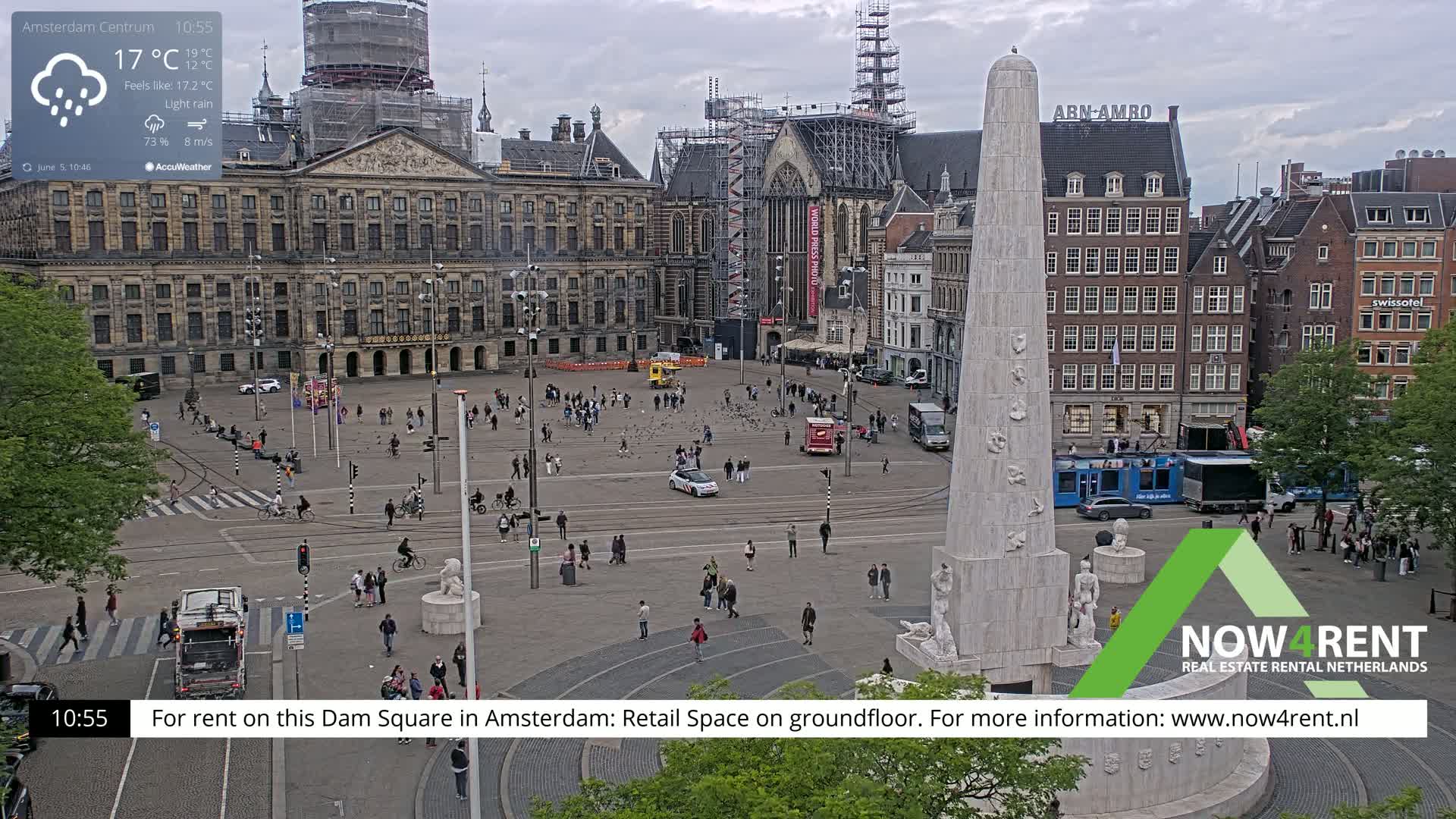 A wide shot of Dam Square in Amsterdam during light rain shows a large monument in the foreground, with people walking around the square and buildings surrounding it, one of which is under construction.