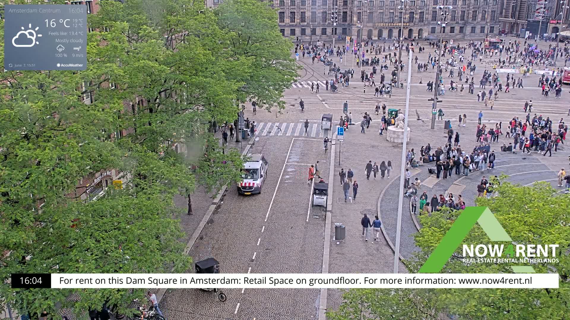 A mostly cloudy, breezy day in a city square with many people, a van, and several buildings visible in the background.