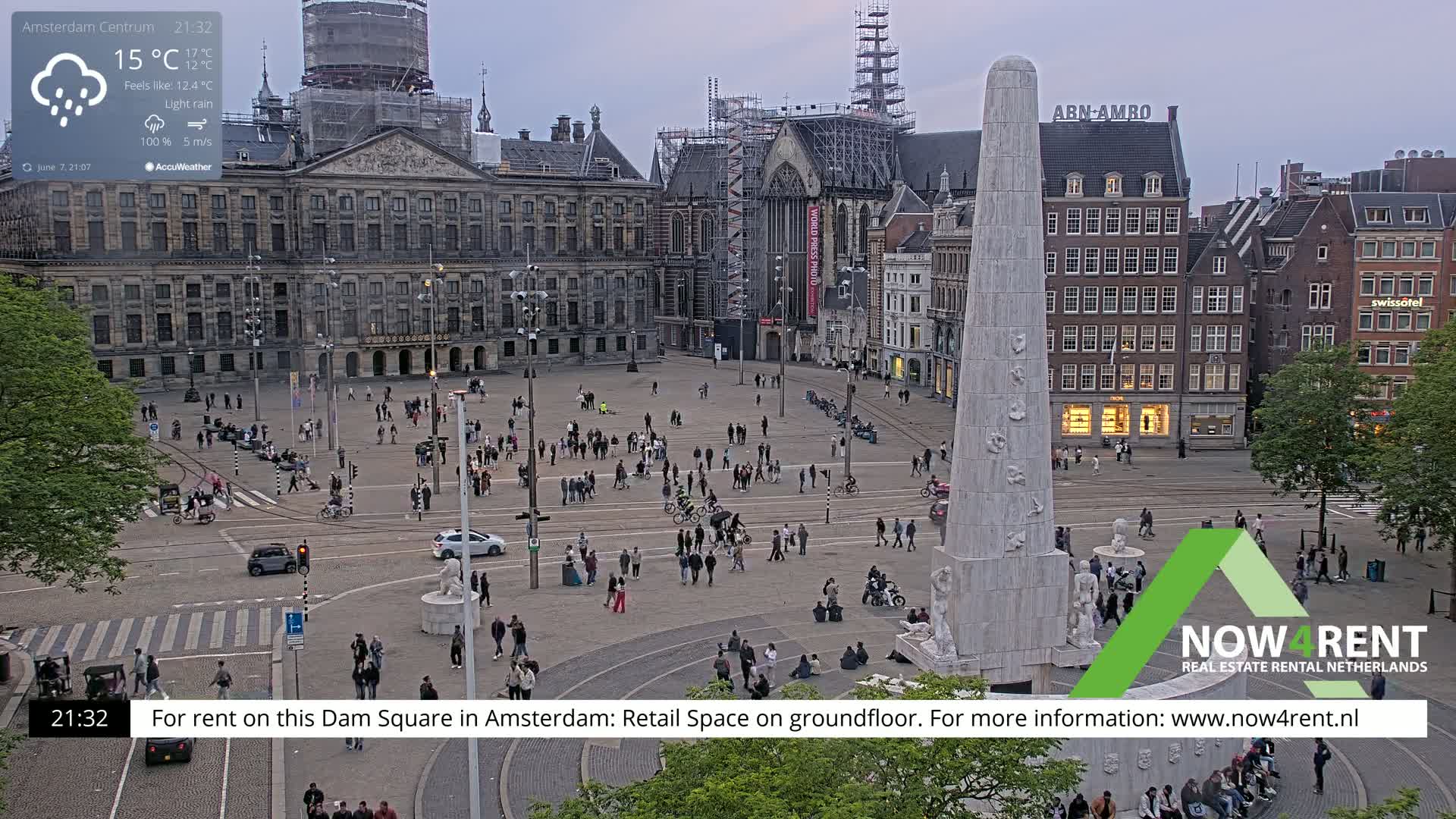 A lightly raining Dam Square in Amsterdam, with many people walking around a central monument and buildings under construction.