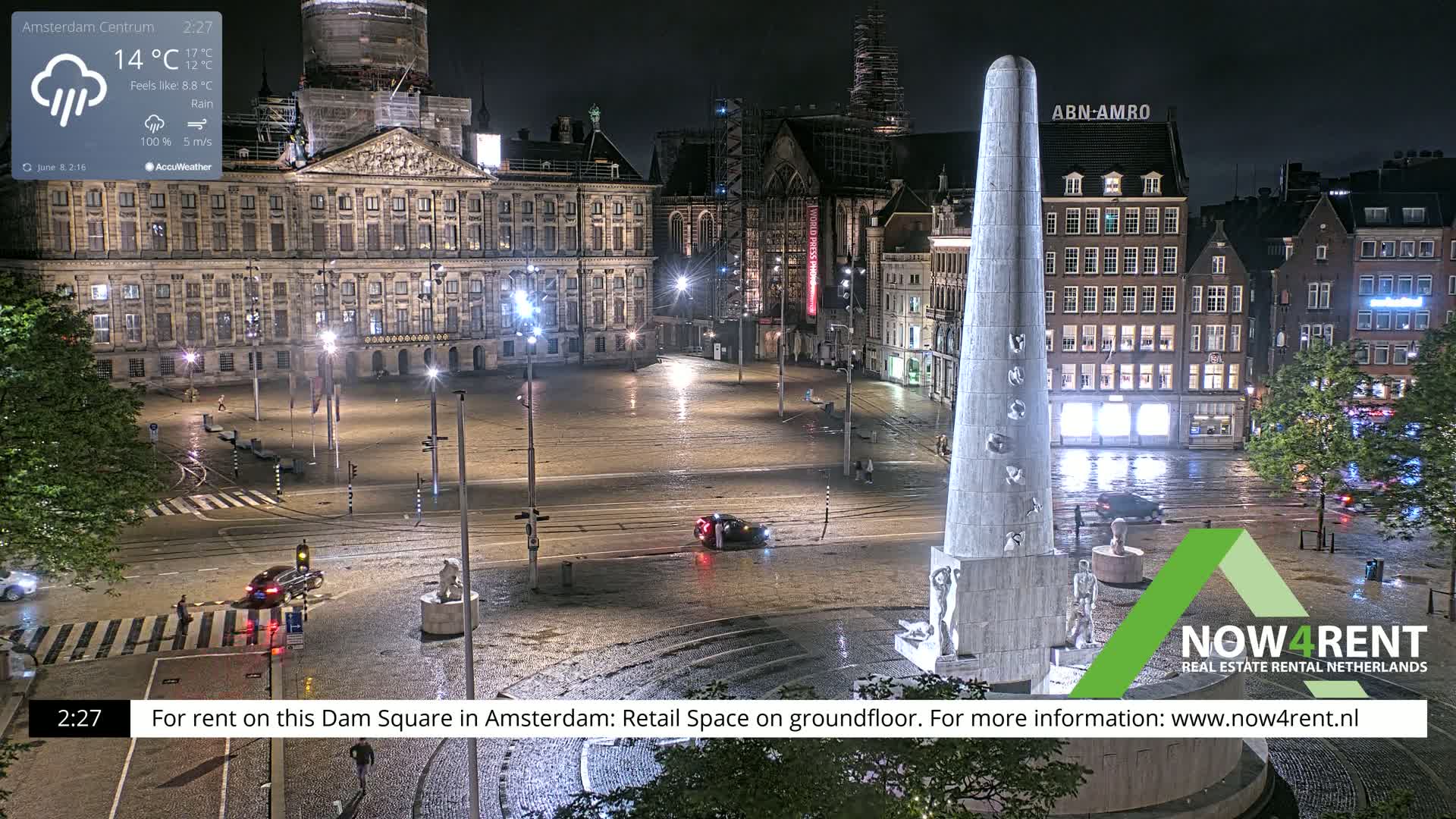 A nighttime view of a rain-slicked Dam Square in Amsterdam, showing a large monument, several buildings, and sparse traffic.