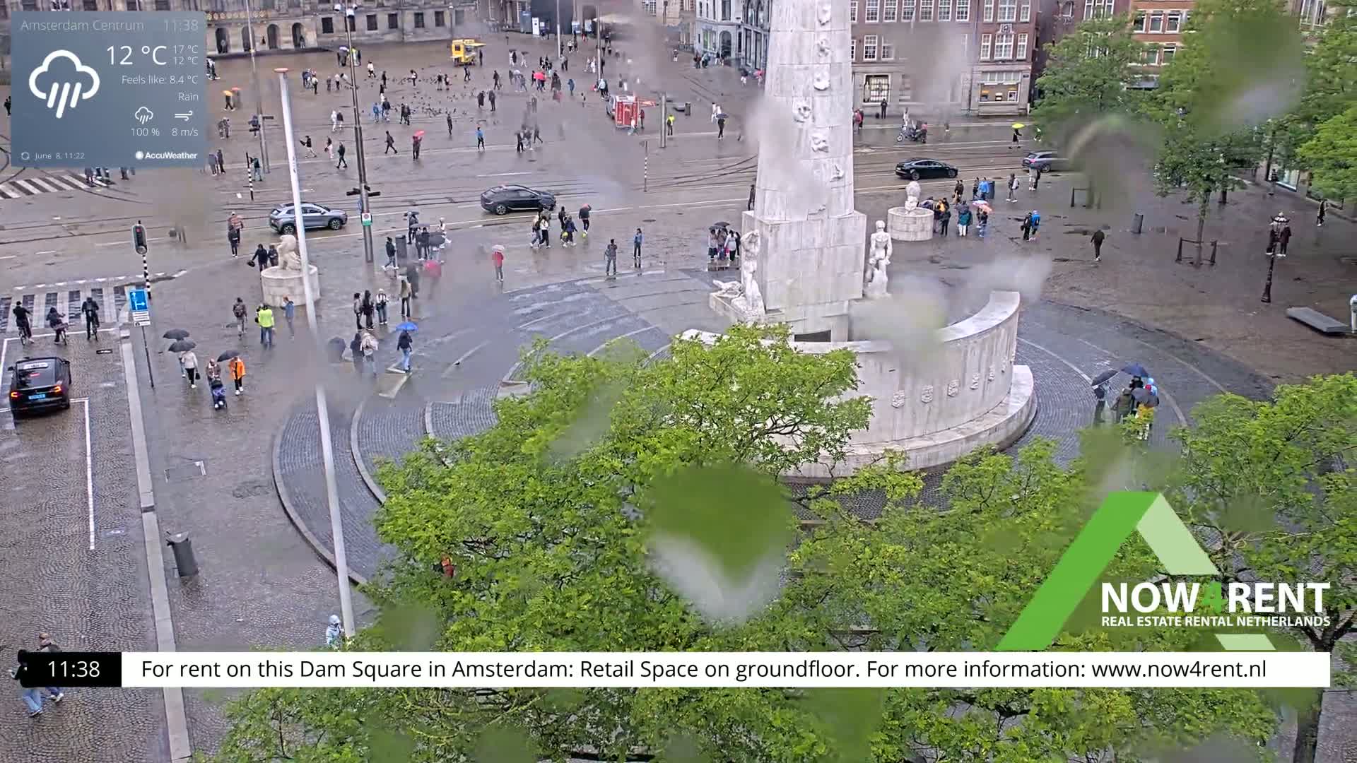 A high-angle, rainy view of a city square with a monument in the center, surrounded by people with umbrellas walking on wet cobblestones.