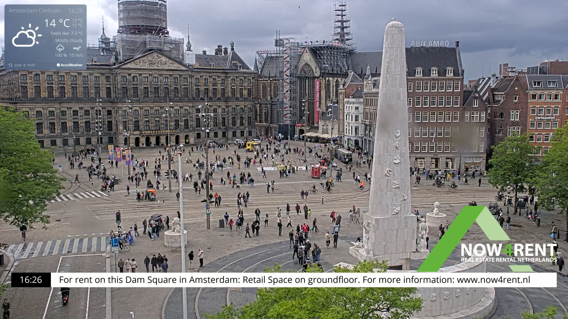 A mostly cloudy day in a large city square, featuring a tall monument surrounded by people and buildings.